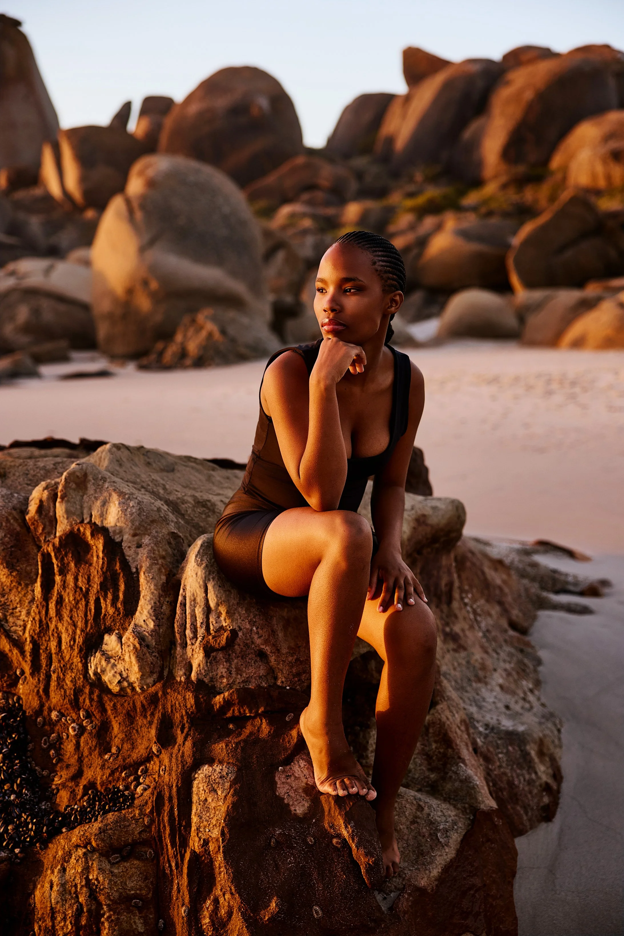 Eine junge Frau sitzt am Meer auf einem Felsen und schaut nachdenklich ins Sonnenlicht