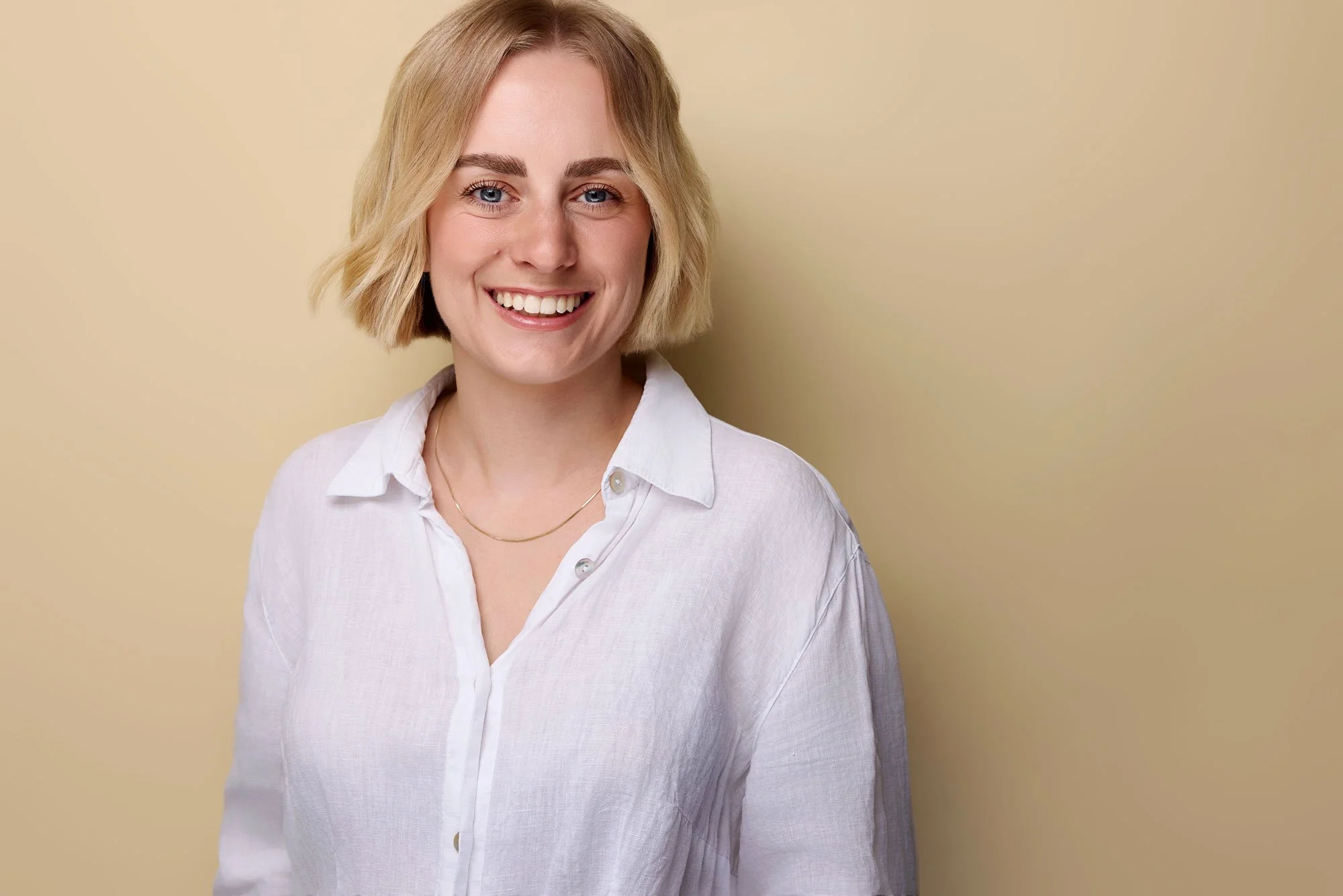 Eine blonde Frau lächelt für ihr LinkedIn Profilfoto in die Kamera des Fotografen und steht vor einer cremefarbenen Wand