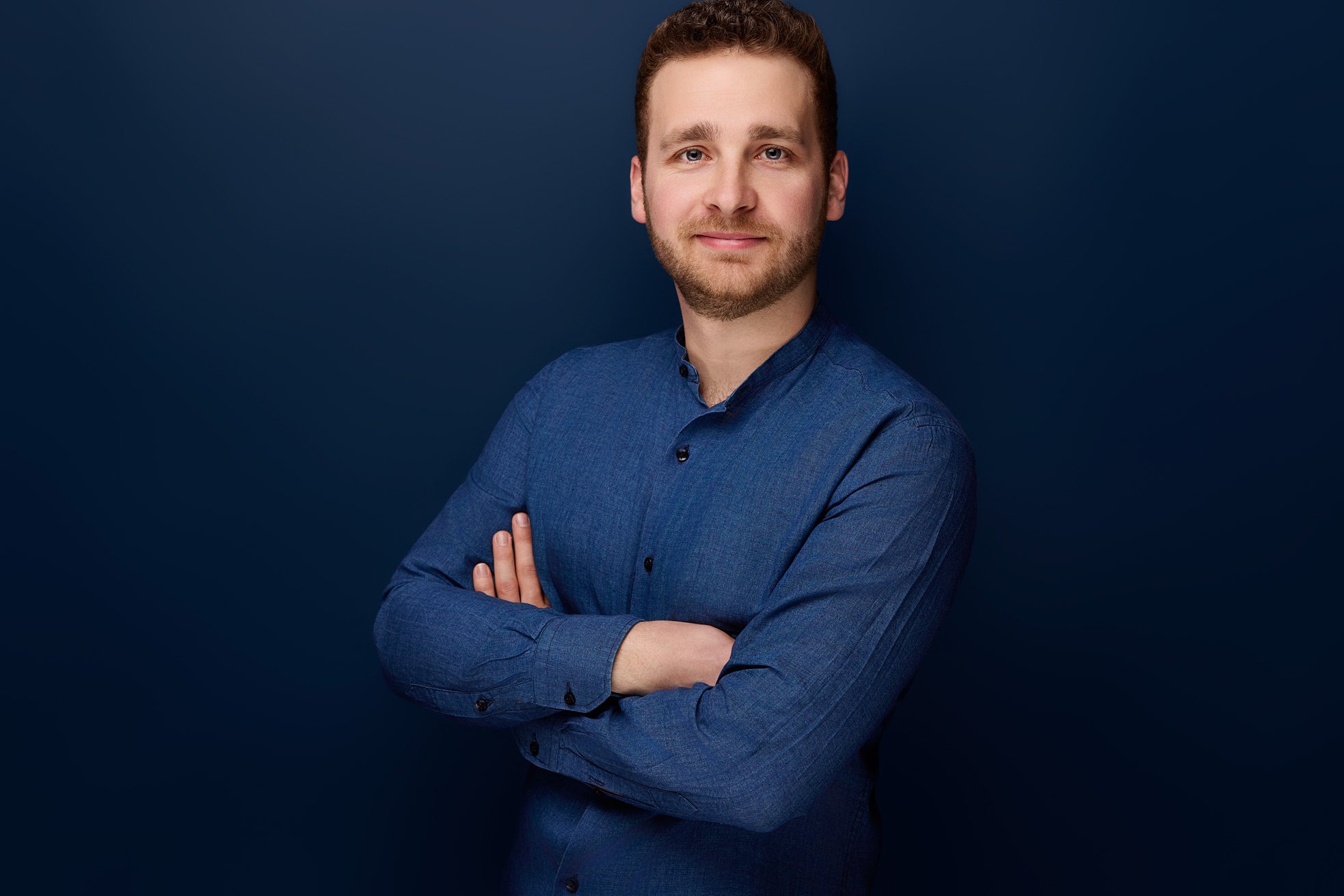 Ein Mann im blauem Hemd steht mit verschränkten armen vor einem blauen Hintergrund