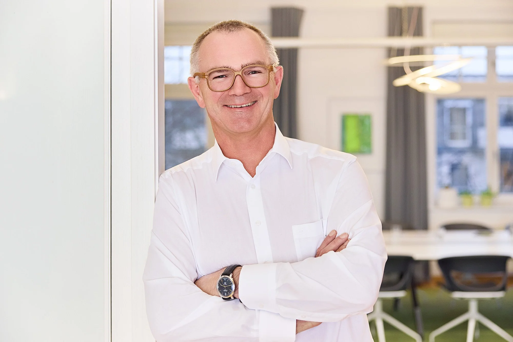 Ein Mann mit Brille und grauen Haaren und weißem Hemd ist am Türrahmen angelehnt in seinem Büro