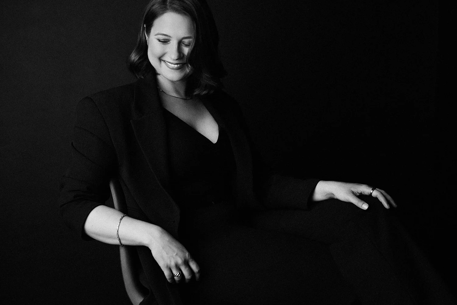 Eine Frau in schwarzen Sachen sitzt lächelnd auf einem Stuhl vor schwarzem Hintergrund