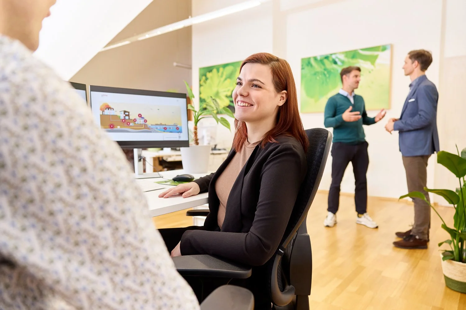 Eine Werbefotografie von einer Frau, die am Rechner sitzt, lächelt zu ihrem Kollegen