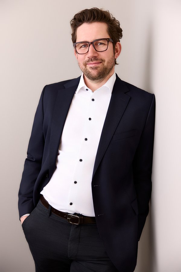 Ein Mann mit dunklen Anzug und weißem Hemd lehnt sich an einer hellgrauen Wand im Fotostudio in Leipzig an