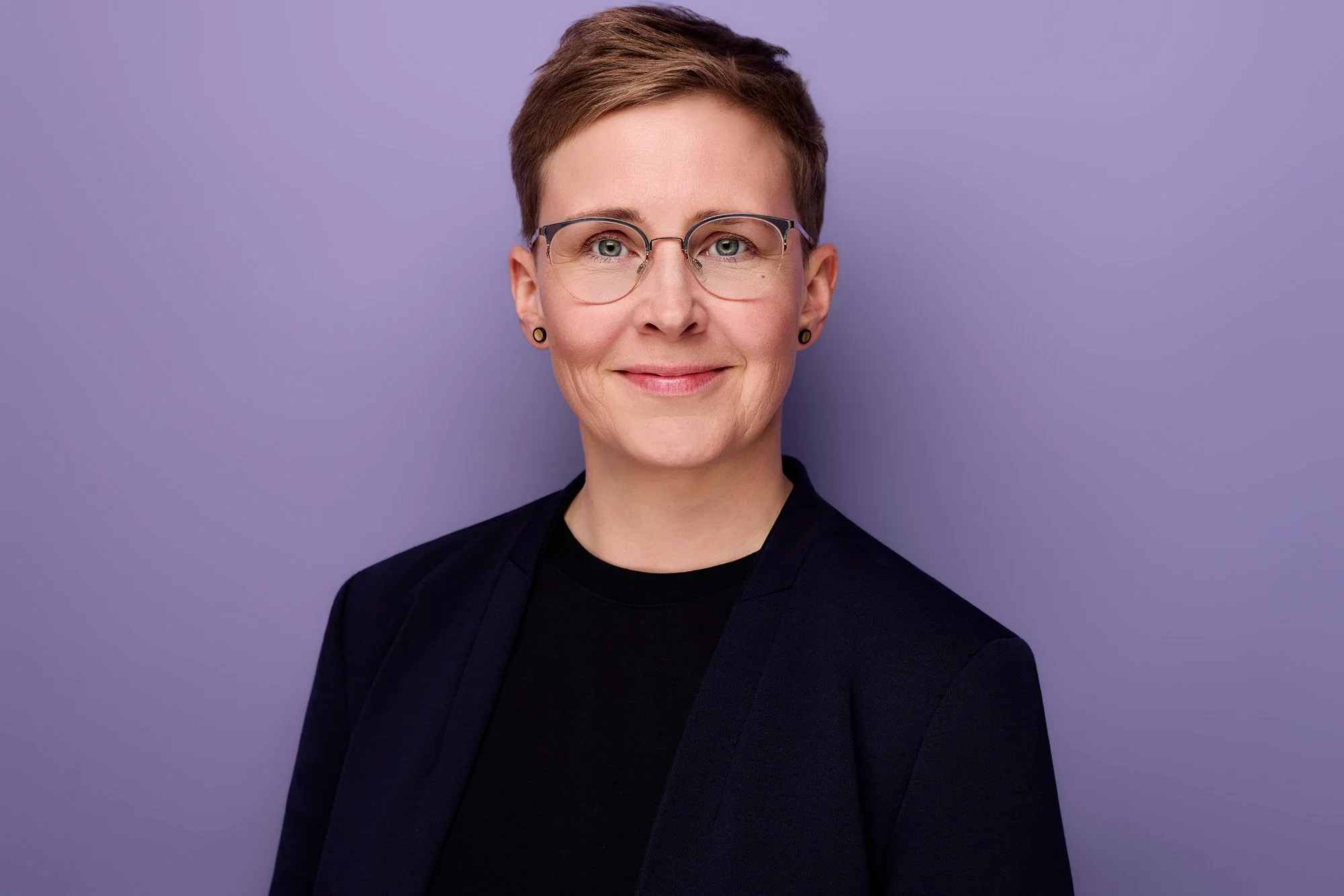 Ein Bewerbungsbild von einer Frau mit dunkler Kleidung vor lilafarbenen Hintergrund