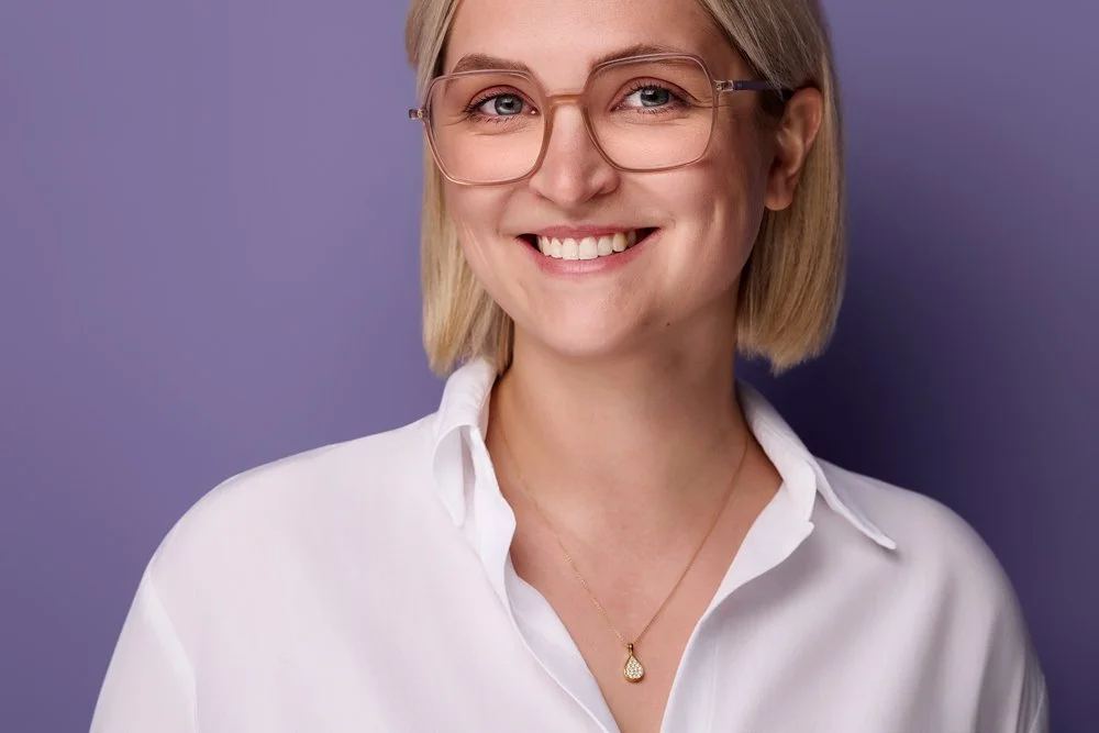 Eine junge Frau mit weißer Bluse und markanter Brille vor einem fliederfarbenen Hintergrund lächelt