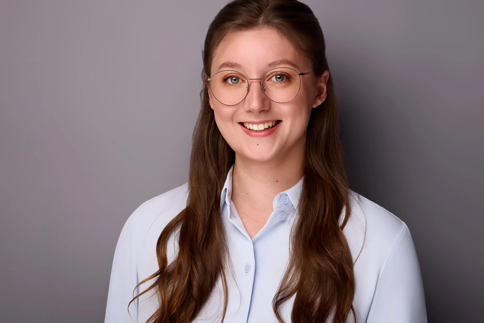 Eine junge Frau mit langen Haaren schaut lächelnd in die Kamera und steht vor einem grauen Hintergrund