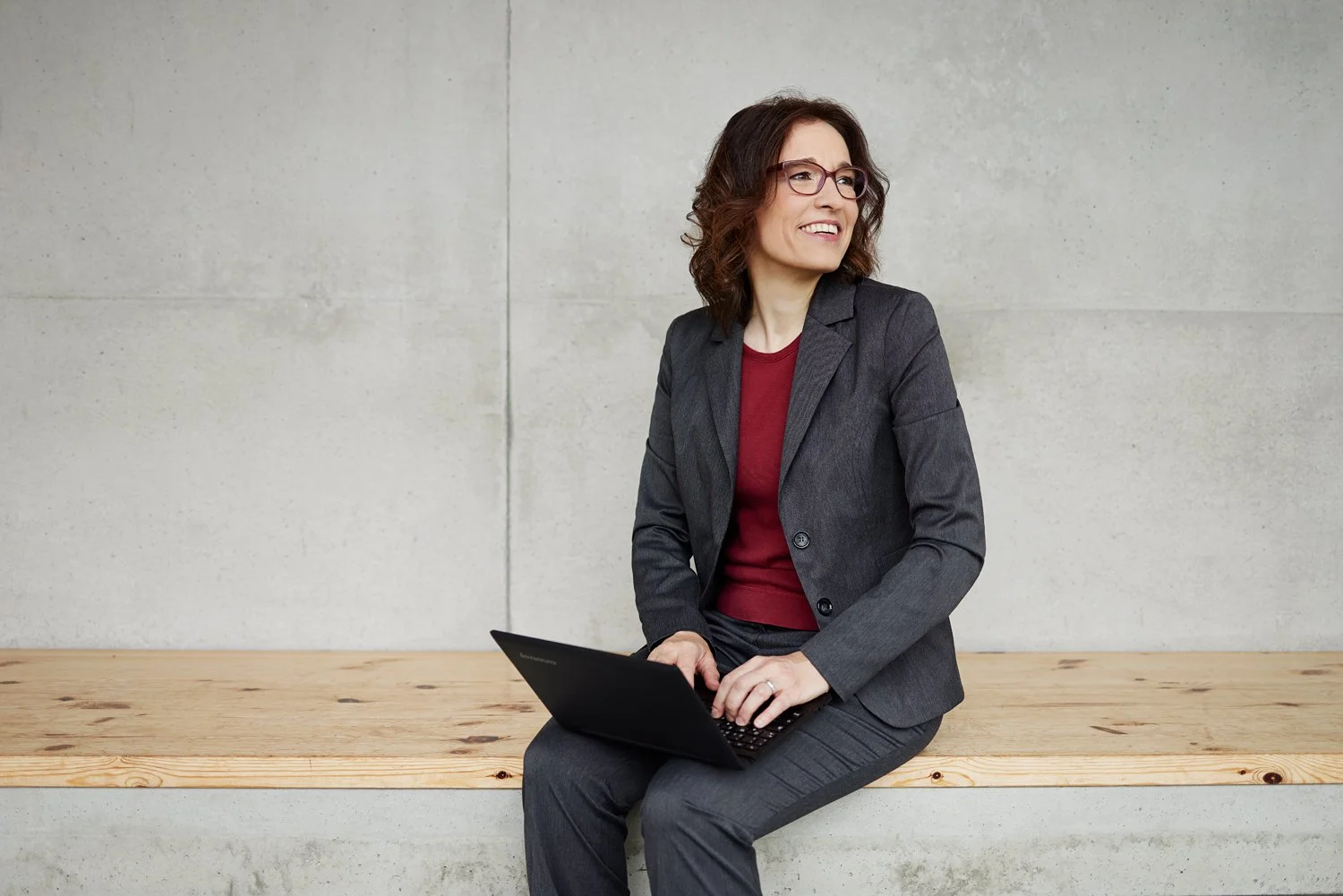 Eine Frau sitzt mit Laptop auf den Knien auf einer Holzbank vor einem Betonhintergrund