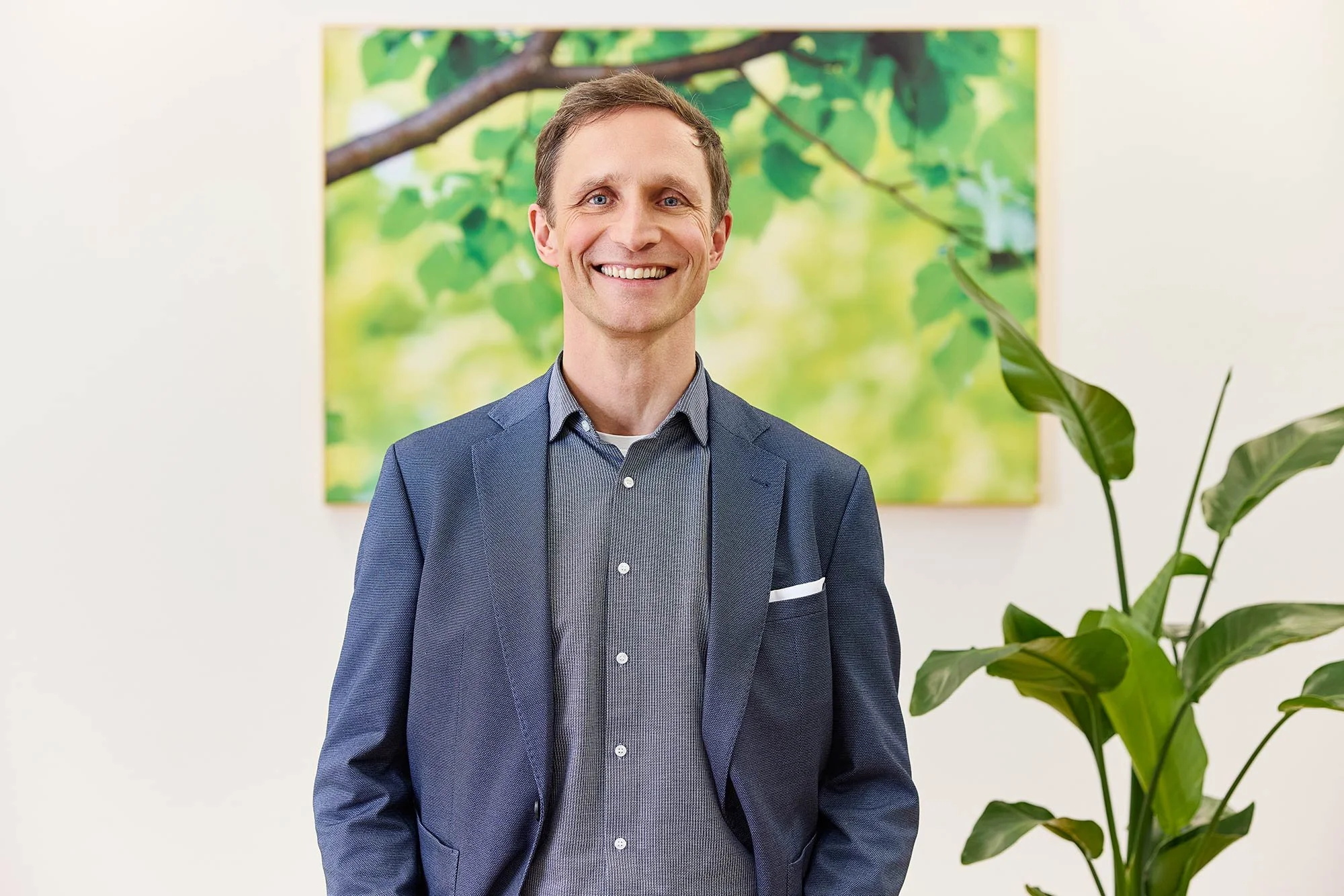 Ein Mann im blauen Sakko steht vor einem grünen Bild und lächelt in die Kamera in seinem Büro