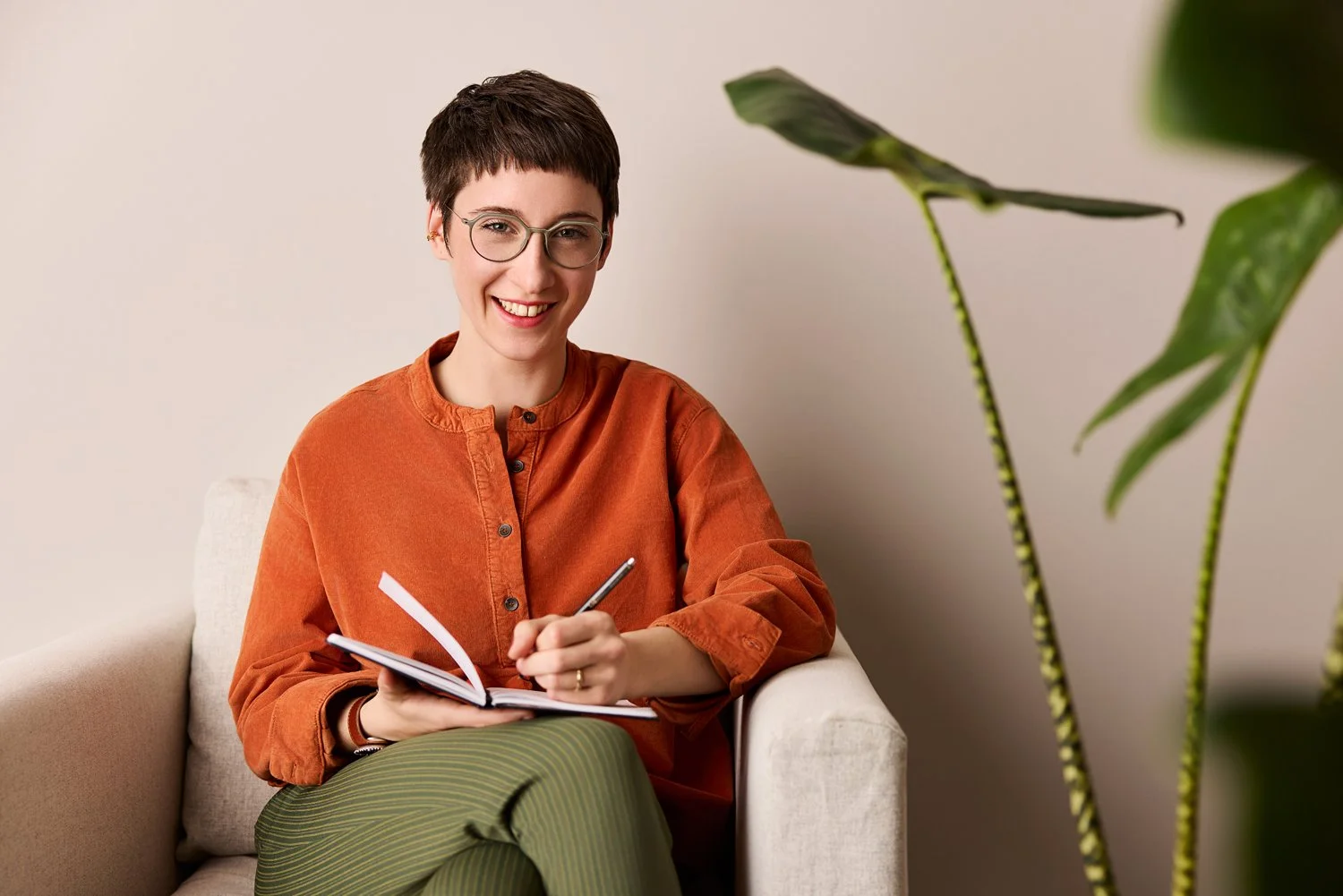 Eine Frau mit markanter Brille sitzt auf einem Sessel und schreibt etwas in ihren Notizbuch, während sie in die Kamera lächelt