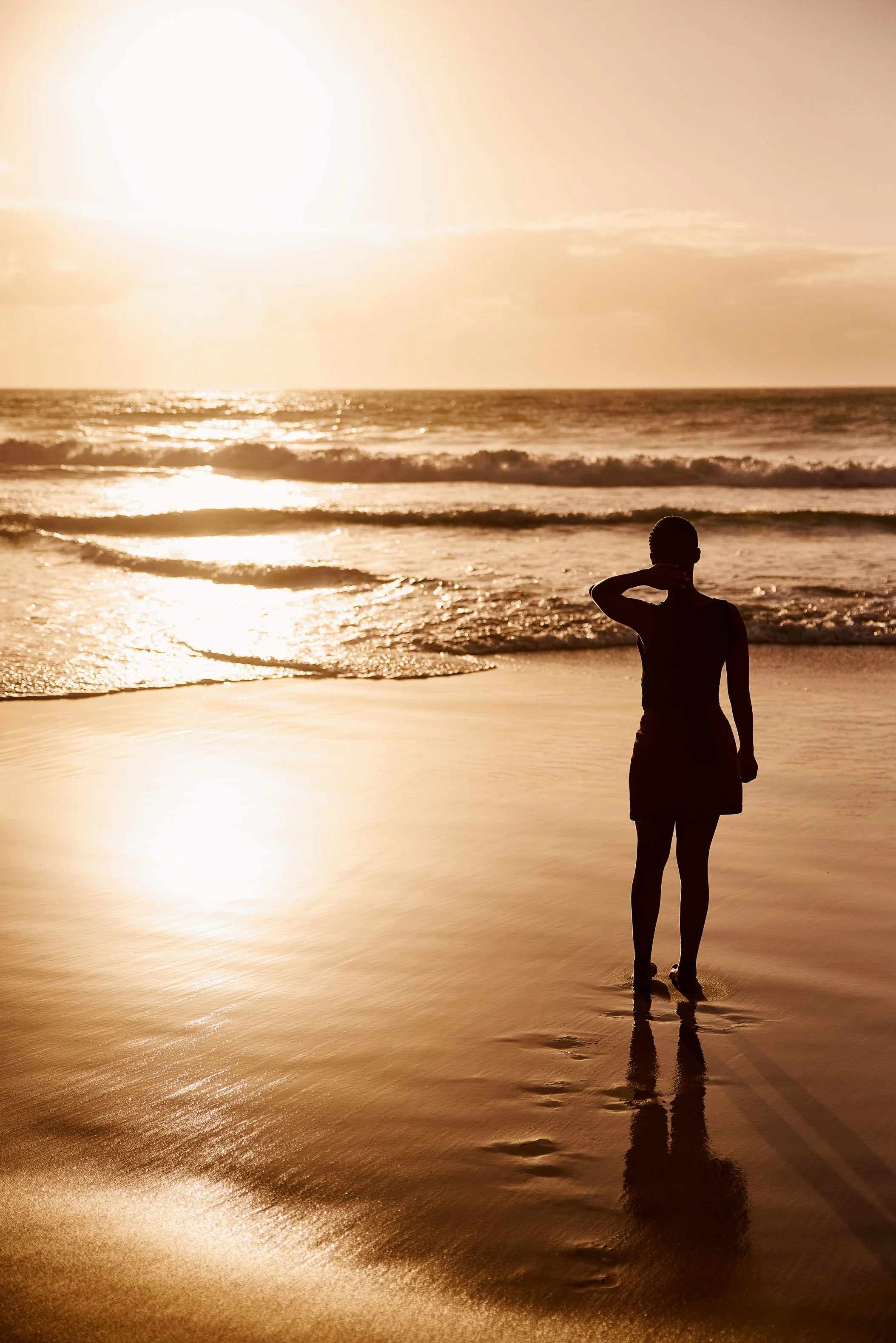 Eine Frau steht barfuß im Sand und schaut hinaus aufs Meer zum Sonnenuntergang