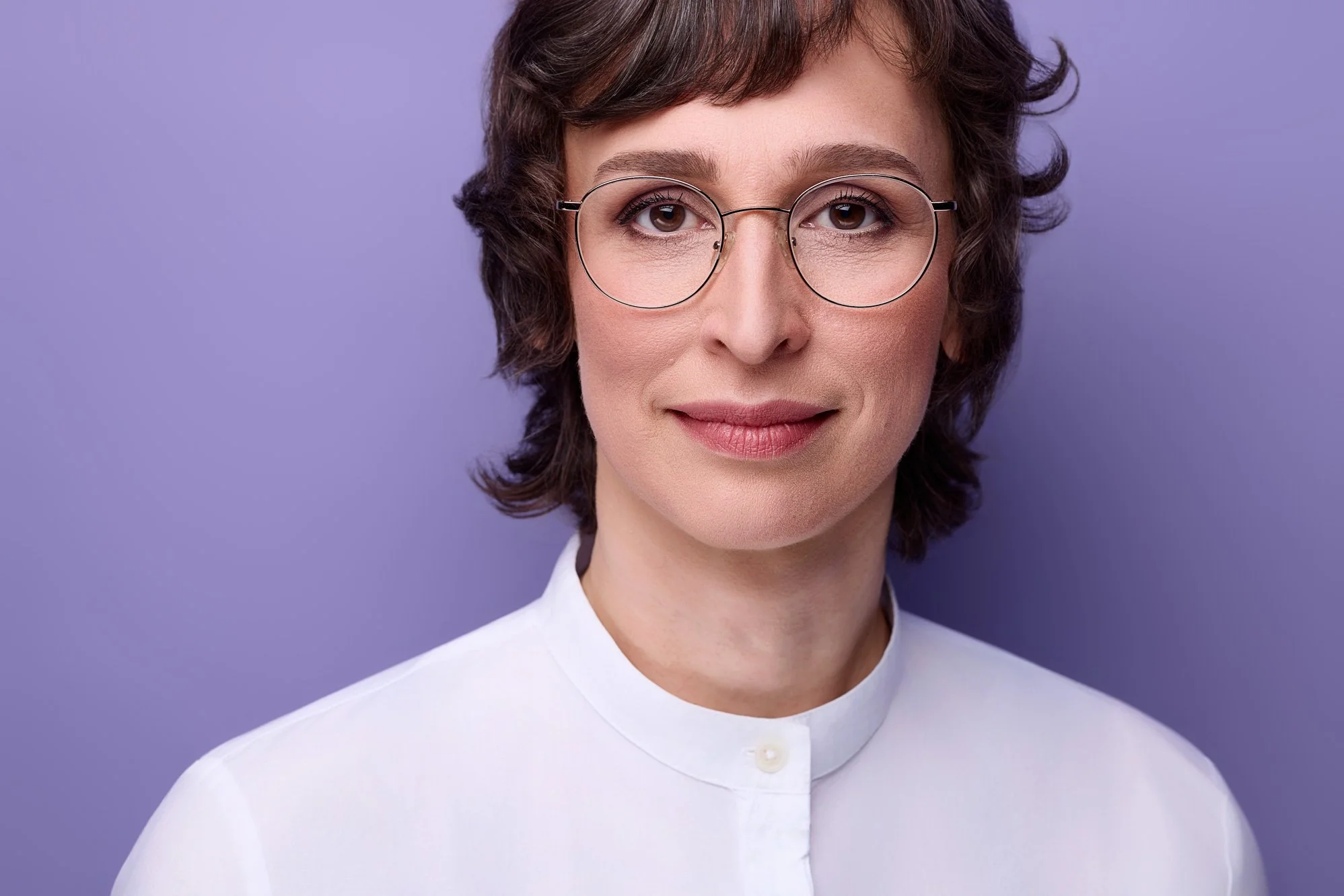 Ein Bewerbungsfoto von einer Frau mit Brille und fliederfarbenen Hintergrund