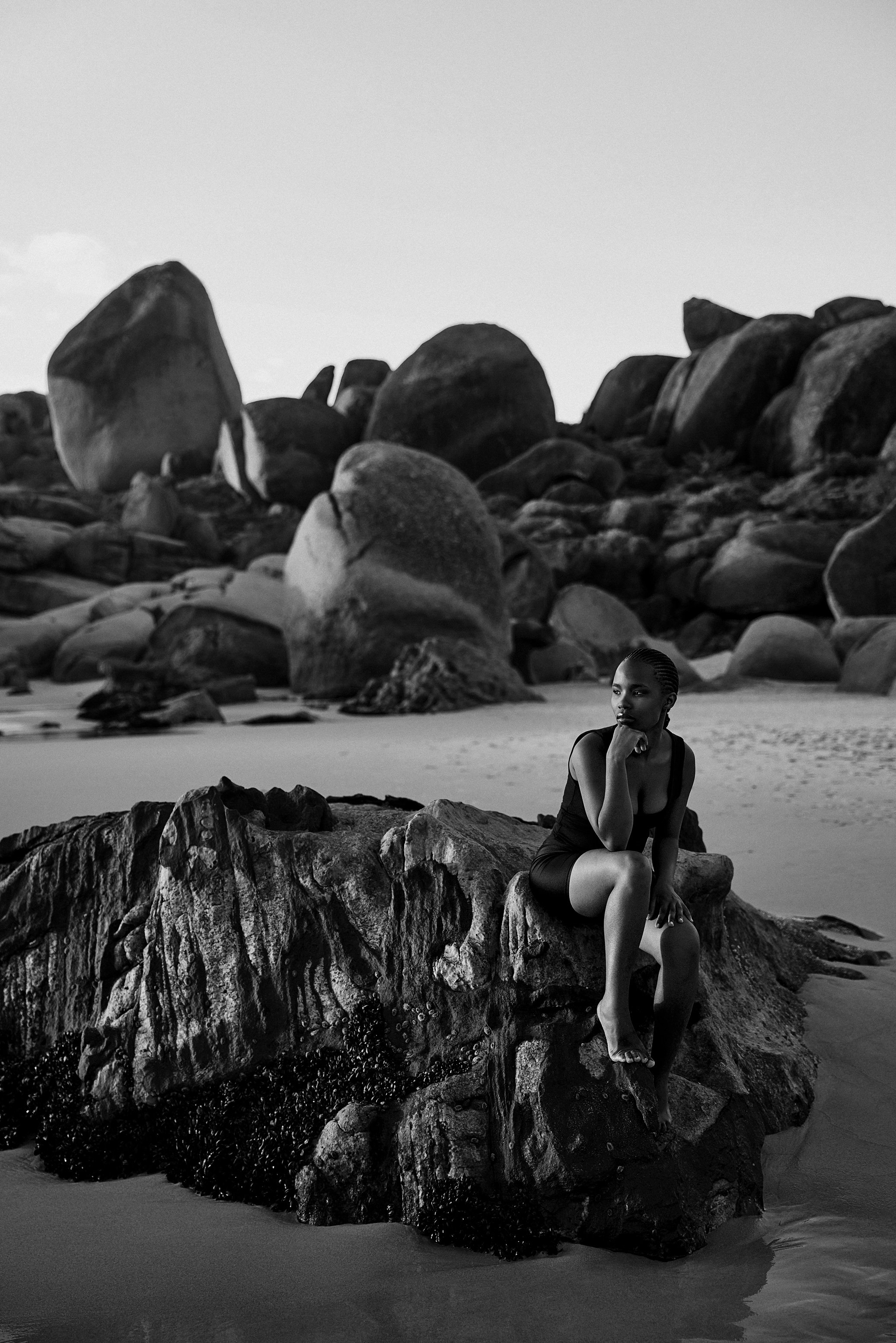 Eine schwarz-weiß Portraitfotografie von einer jungen Frau, die am Strand auf einem Felsen sitzt