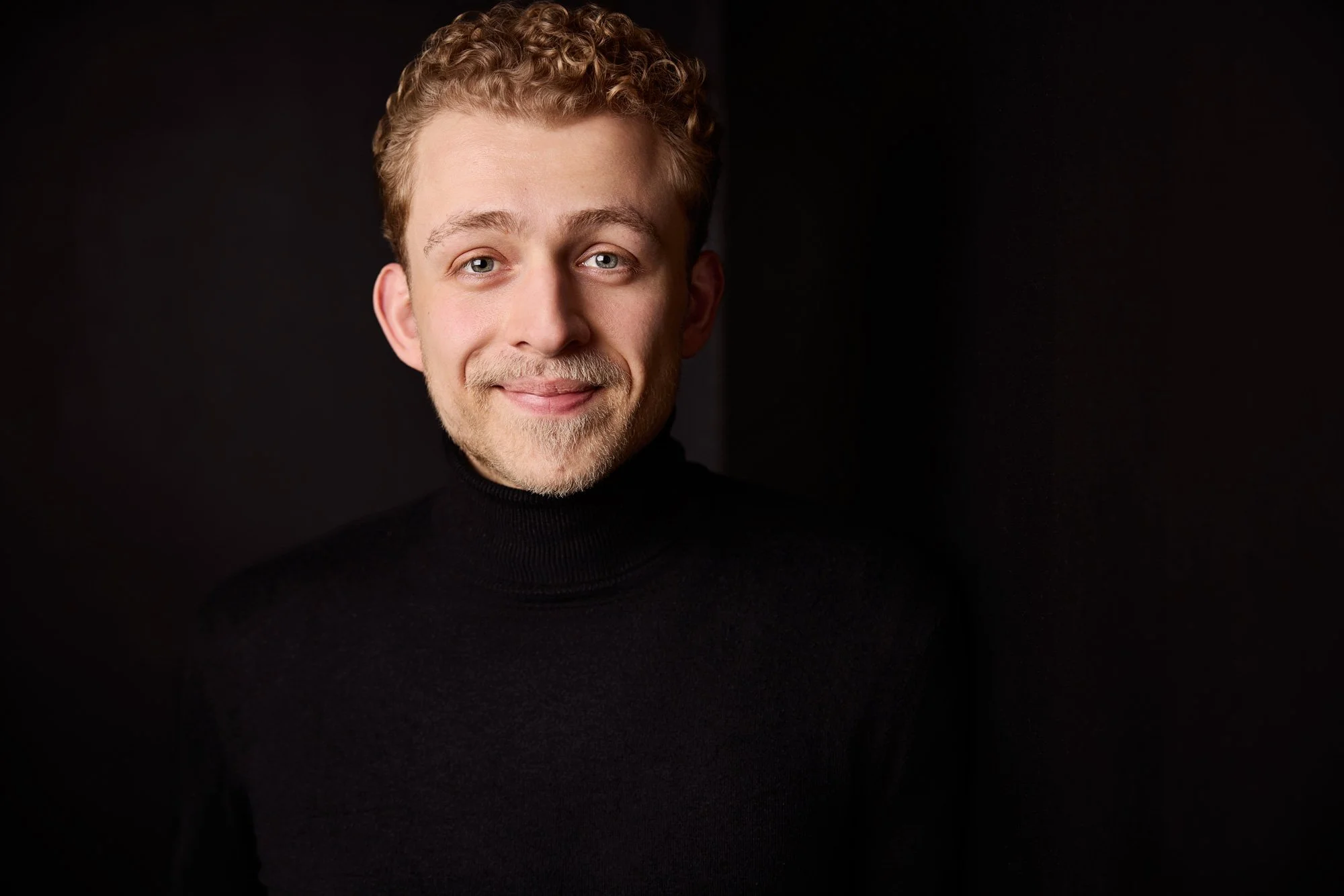 Ein junger Mann lächelt in die Kamera, vor einem schwarzen Hintergrund und schwarz gekleidet