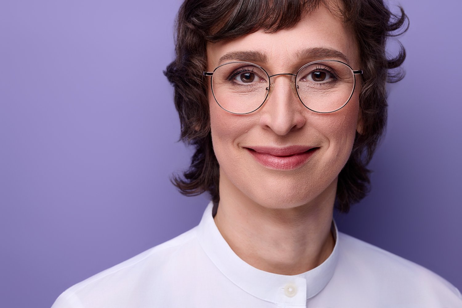 Eine Frau mit dunklen Haaren und Brille auf einem Bewerbungsfoto mit Fliederfarben im Hintergrund