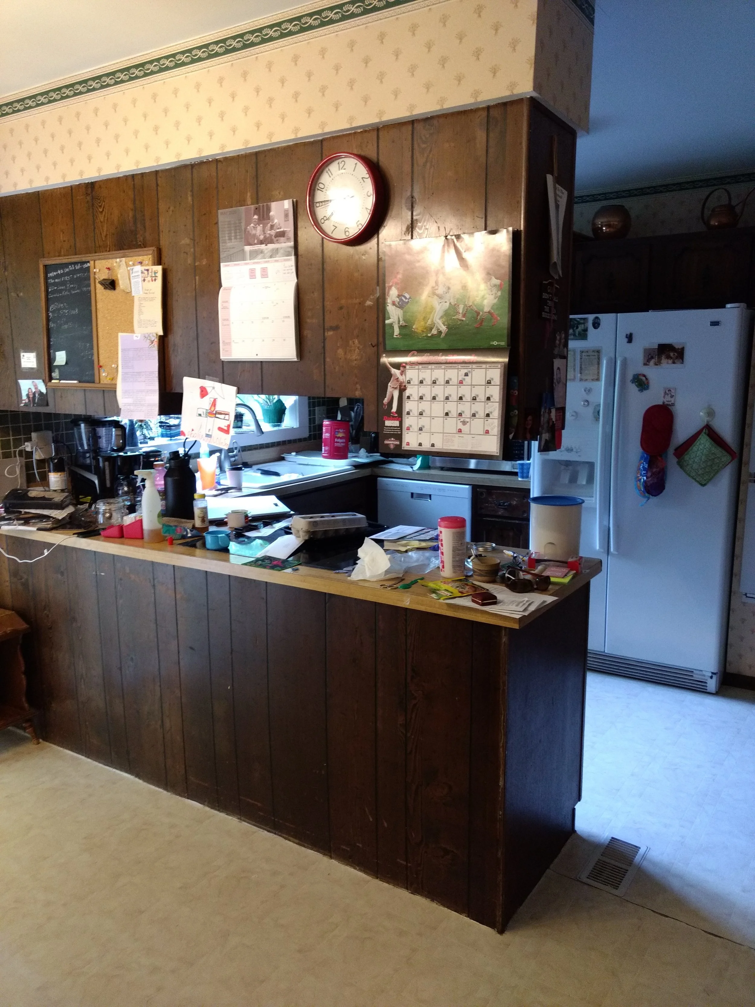 Before. Dark wood paneling and cabinets, wallpaper and a cramped space with a u-shape kitchen.