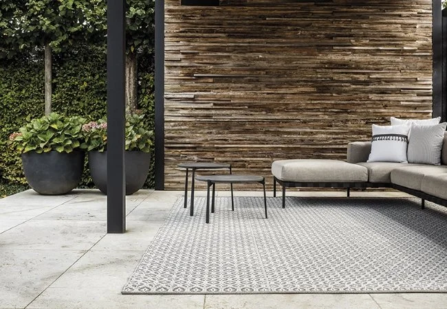 Une terrasse extérieure avec un canapé beige, des tables basses, un tapis gris, des plantes en pot, et un panneau en bois.
