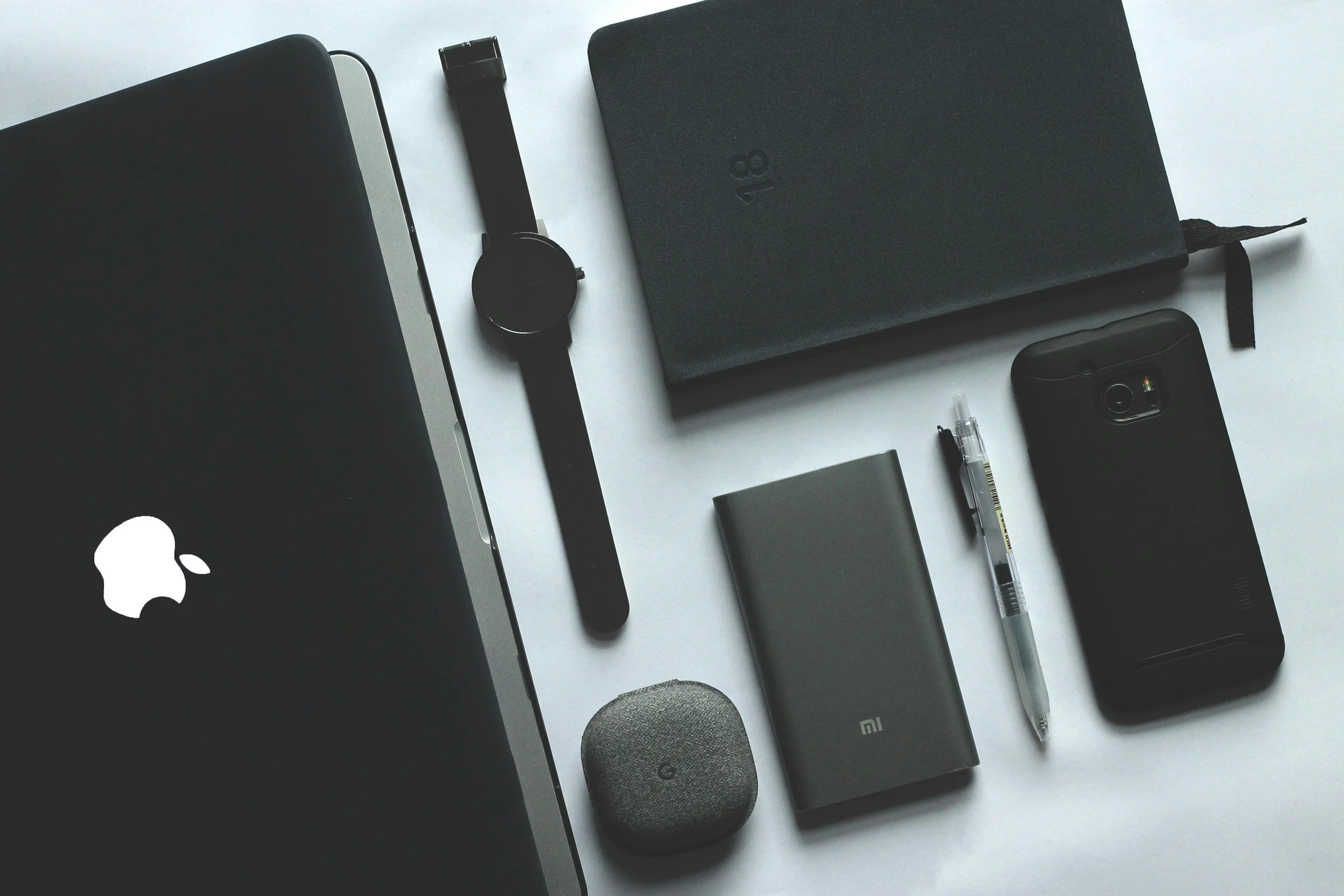 Arrangement of modern tech gadgets on a white surface, including a black Apple MacBook, black smartwatch, black notebook, black smartphone, gray power bank, black portable speaker, clear pen, and a black pen.