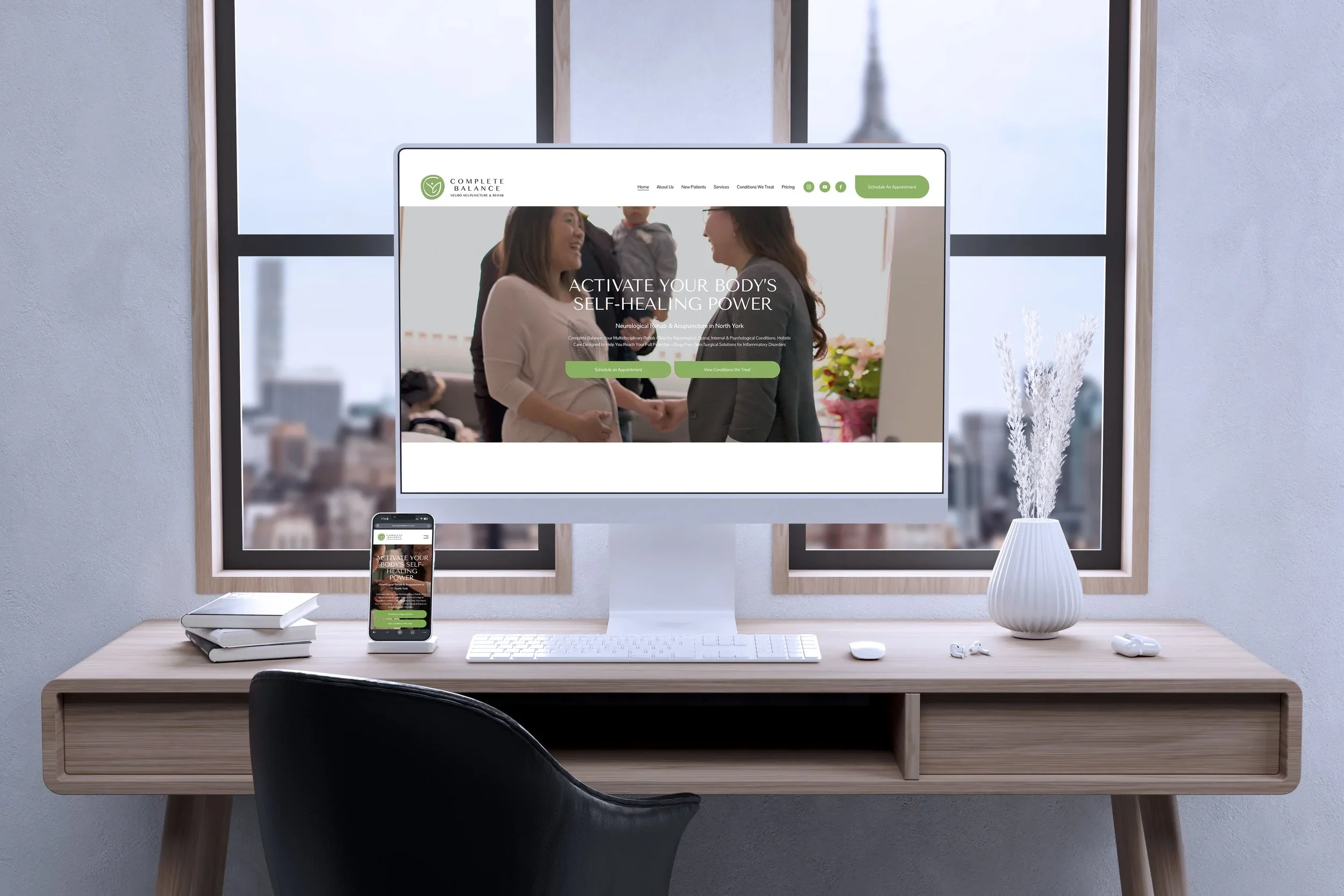 Computer monitor, smartphone, keyboard, mouse, and a coffee table on a desk with a window in the background. The monitor displays a website for Complete Balance, a neuro acupuncture and rehab clinic, showing an image of two women shaking hands with a child, and text promoting self-healing power.
