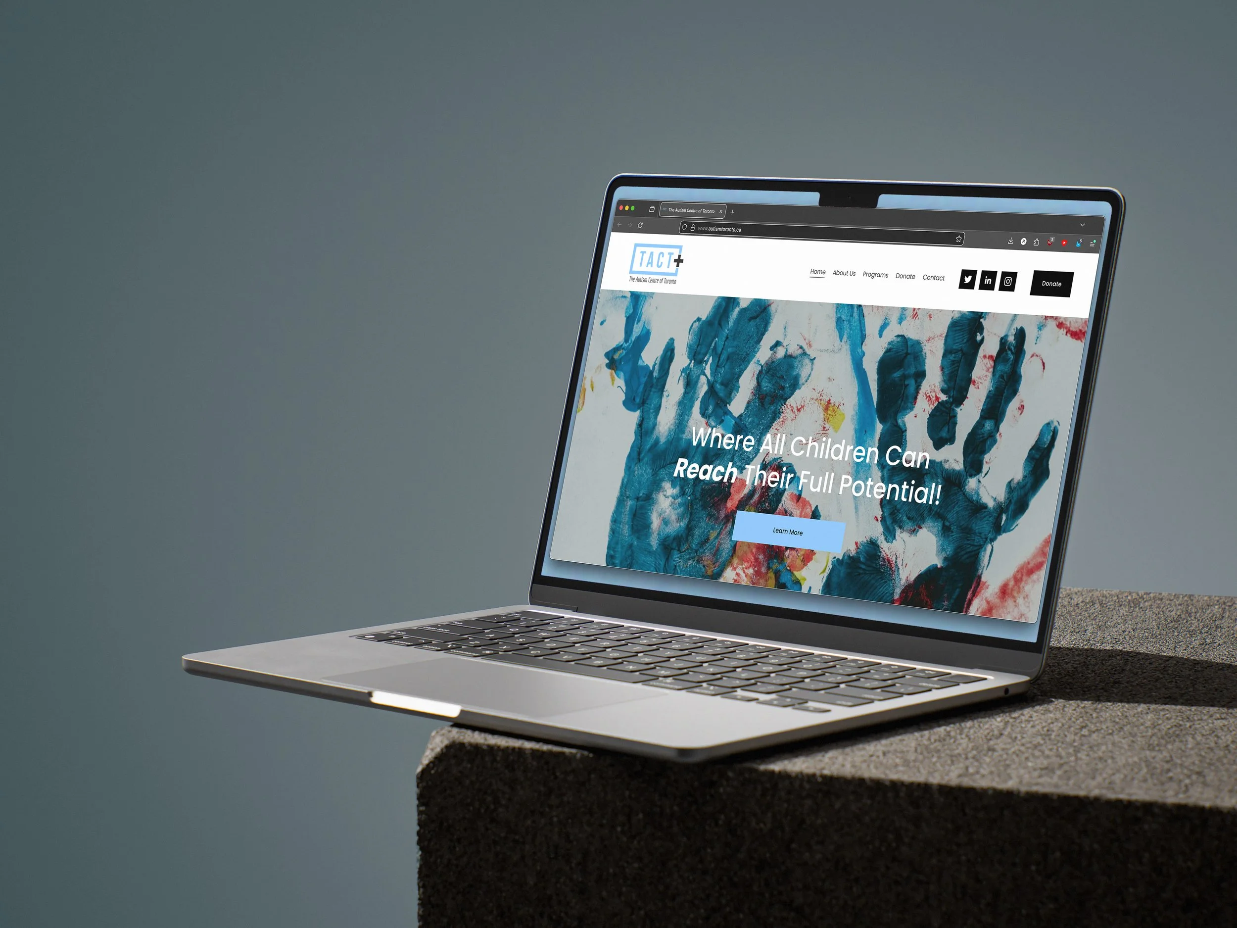 A laptop computer displaying a website with a colorful handprint art background, placed on a concrete surface outdoors.