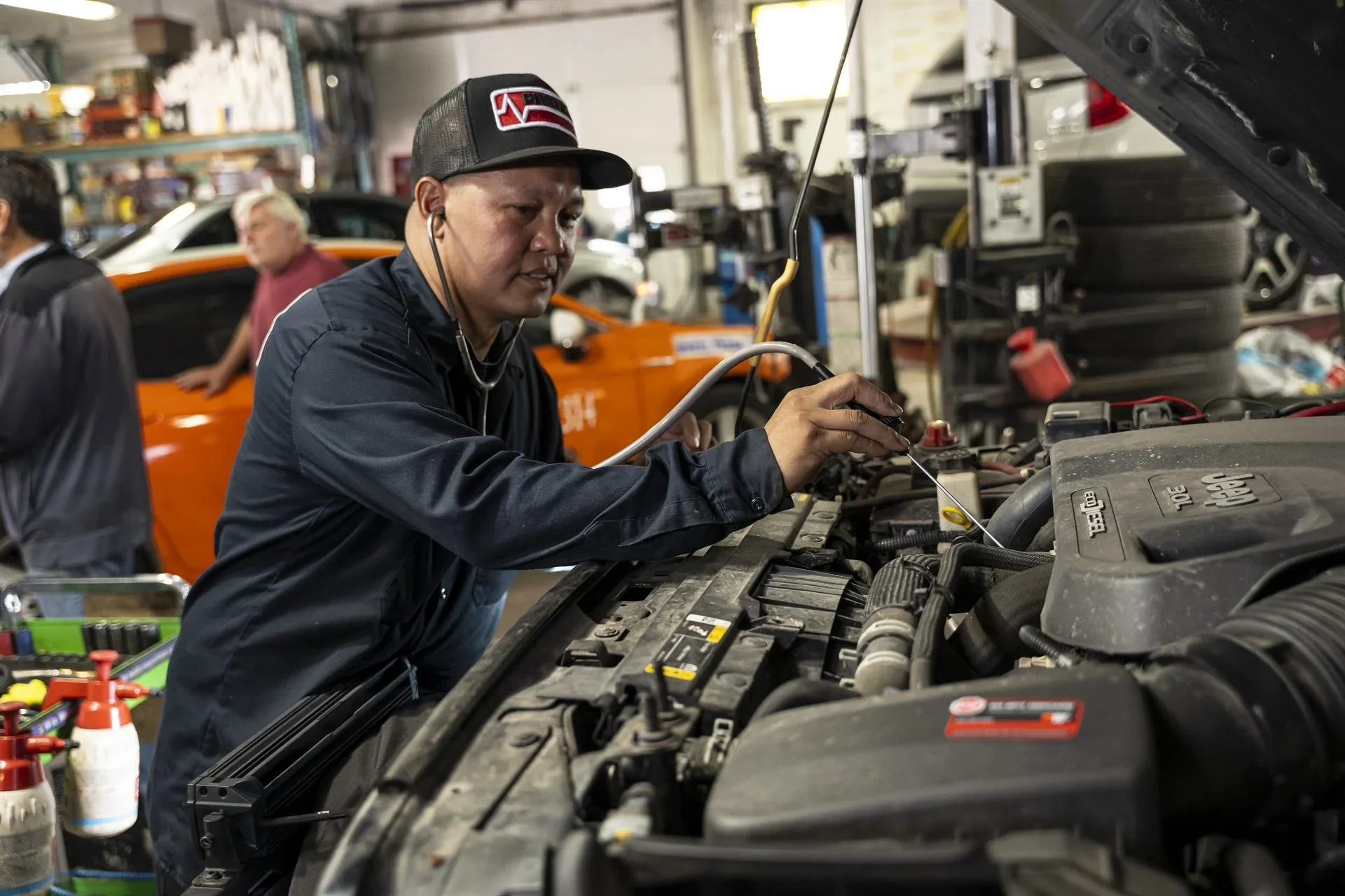 A mechanic wearing a cap and a black jacket is inspecting the engine of a vehicle in an auto repair shop, with tools and a bright orange car in the background.