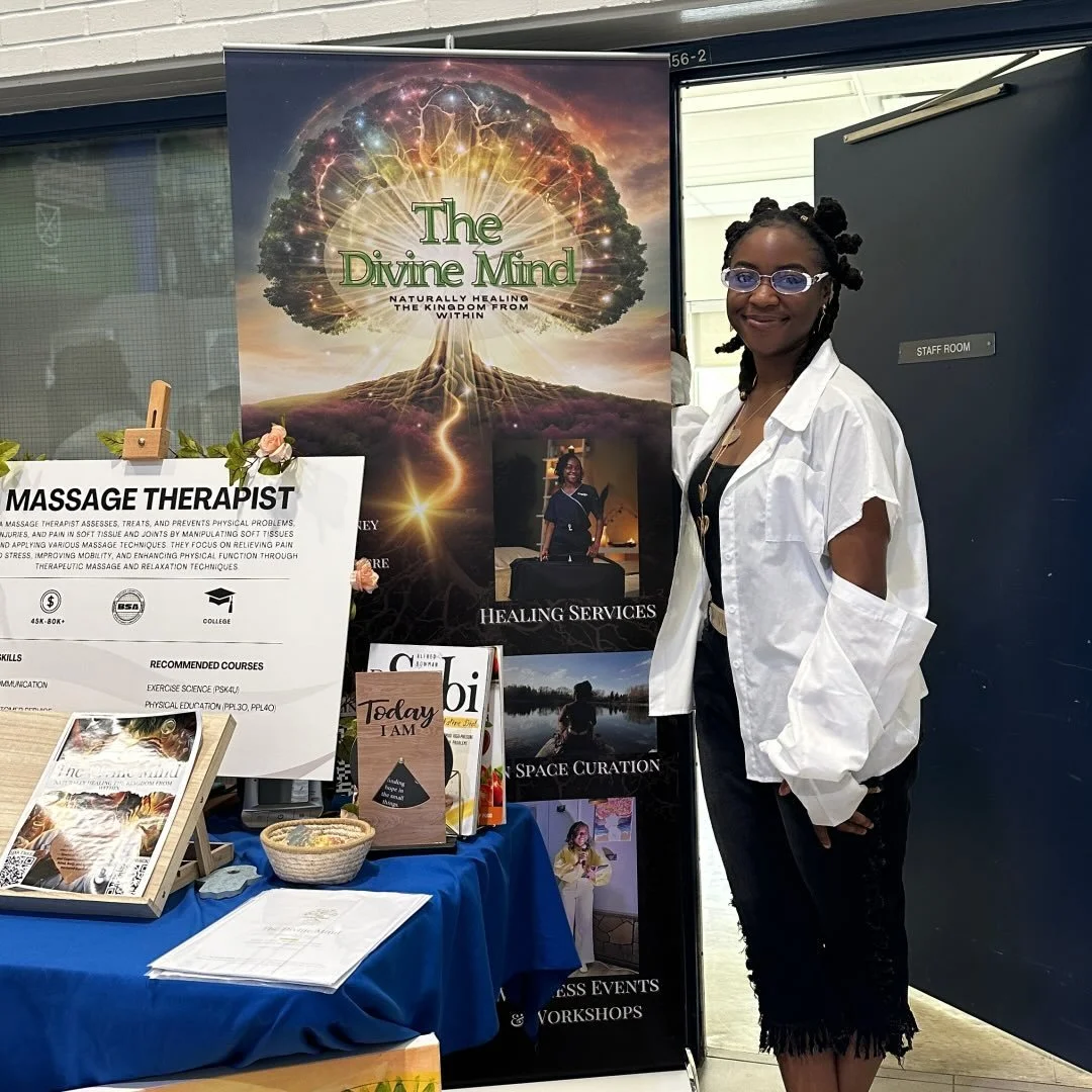 A woman standing next to a booth featuring a large poster for 'The Divine Mind' with a glowing brain-shaped tree and lightning. The booth promotes healing and space curation services, with informational signs and pamphlets on the table.