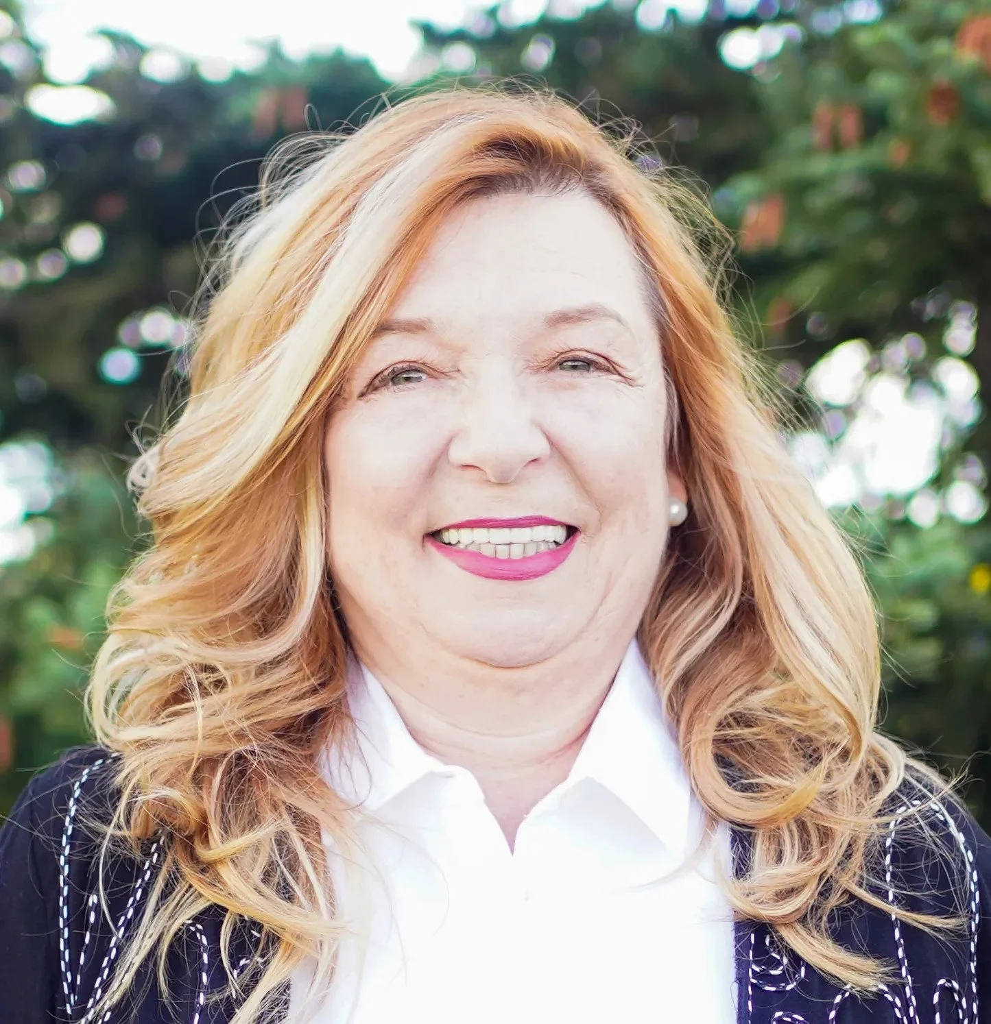 A woman with curly blonde hair smiling outdoors, wearing a white collared shirt and a dark blazer with white stitching, against blurred green foliage background.