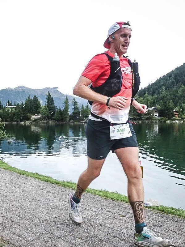 Male ultra marathon runner, running next to a lake. He is wearing a red shirt with a black running pack, black shorts, running shoes, and has maori tattoos on his lower legs. The background shows a clear  sky with mountains covered in trees.