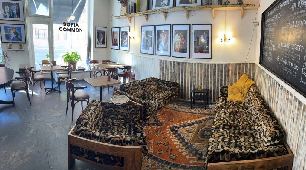 Inomhusmiljö av ett café eller ett litet restaurang. Det finns flera bord och stolar, väggbilder, en svart tavla med text och en sittgrupp med sammetsklädda soffor med ett mönster, en matta och en liten bord. Fönster med gröna växter utanför. Sol- el