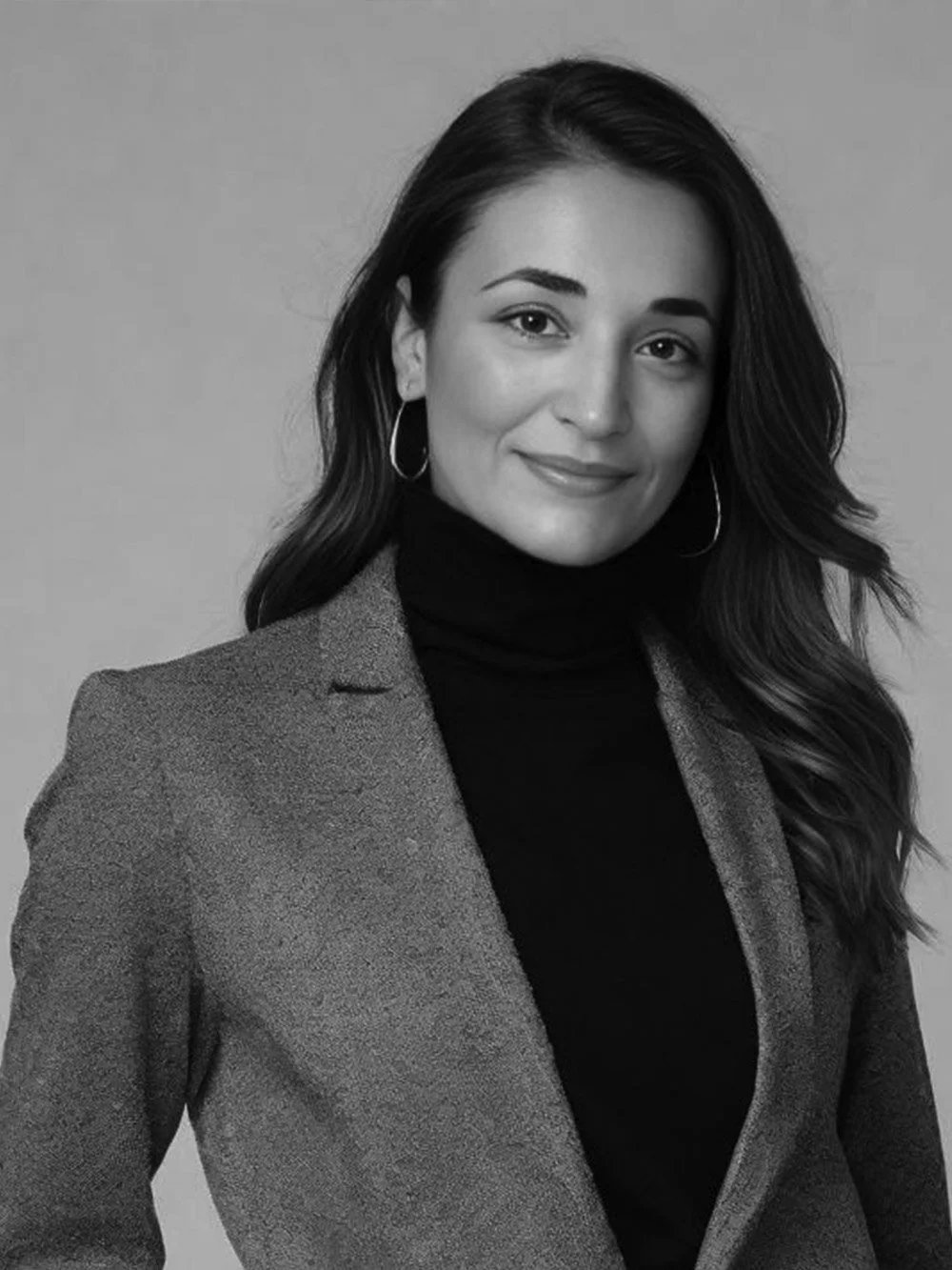 Black and white portrait of a woman with long, wavy hair wearing hoop earrings, a dark turtleneck, and a blazer.