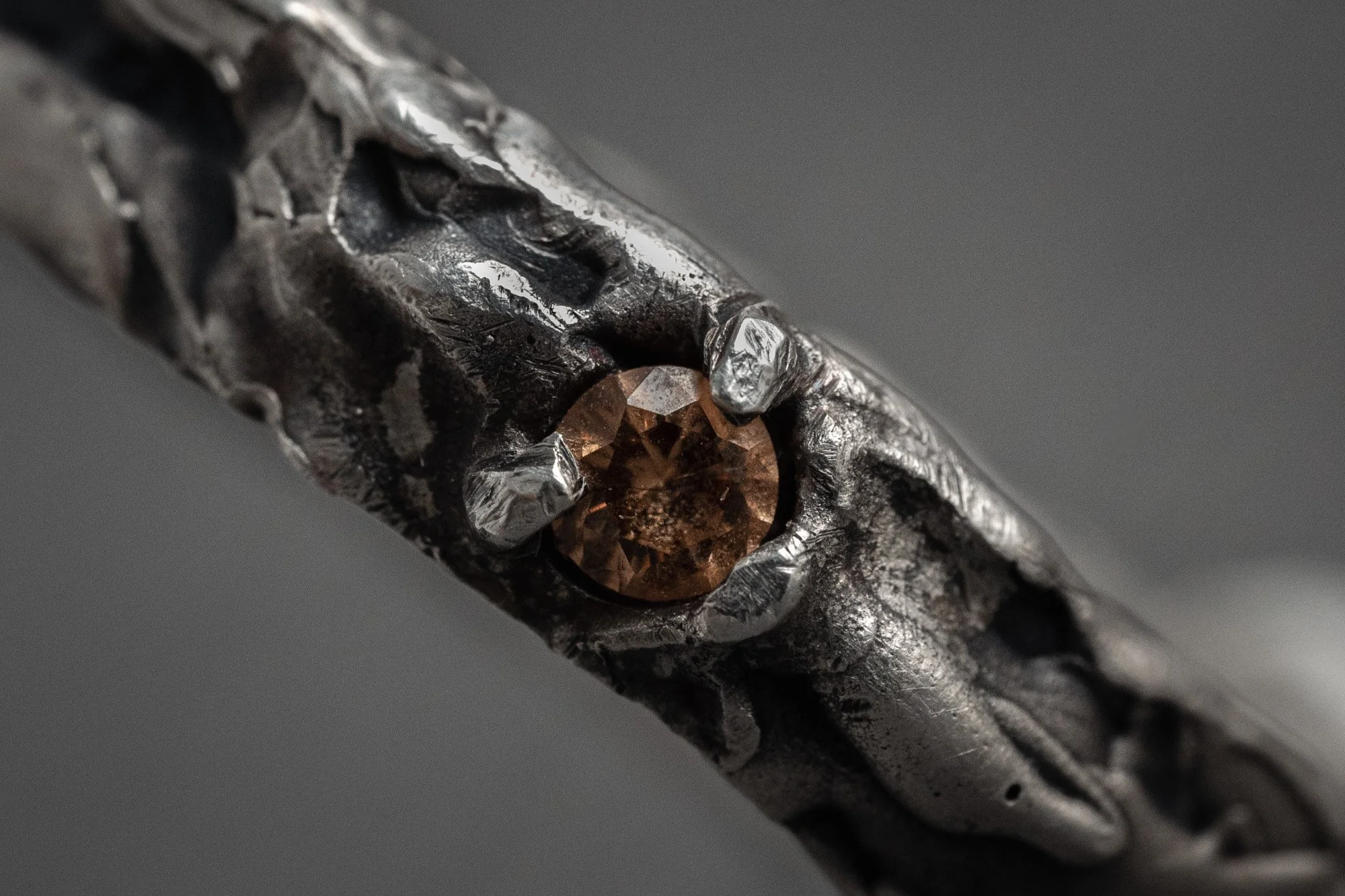 Primer plano de un anillo de metal con un engaste que sostiene una piedra preciosa marrón. El diseño del metal es rugoso y texturizado.