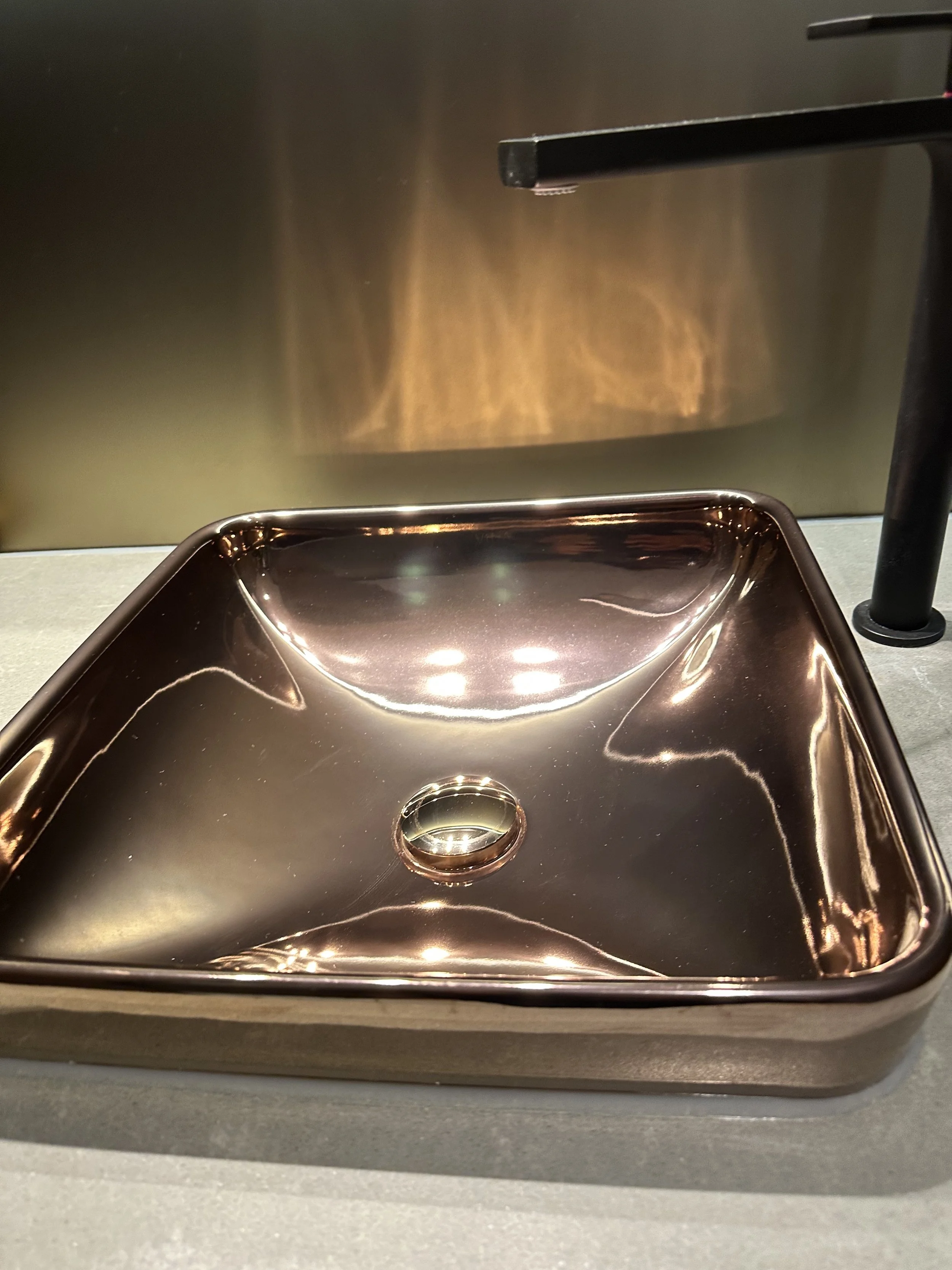 A shiny metallic copper-colored sink with a round drain hole in the center, set against a dark wall with reflected lighting.