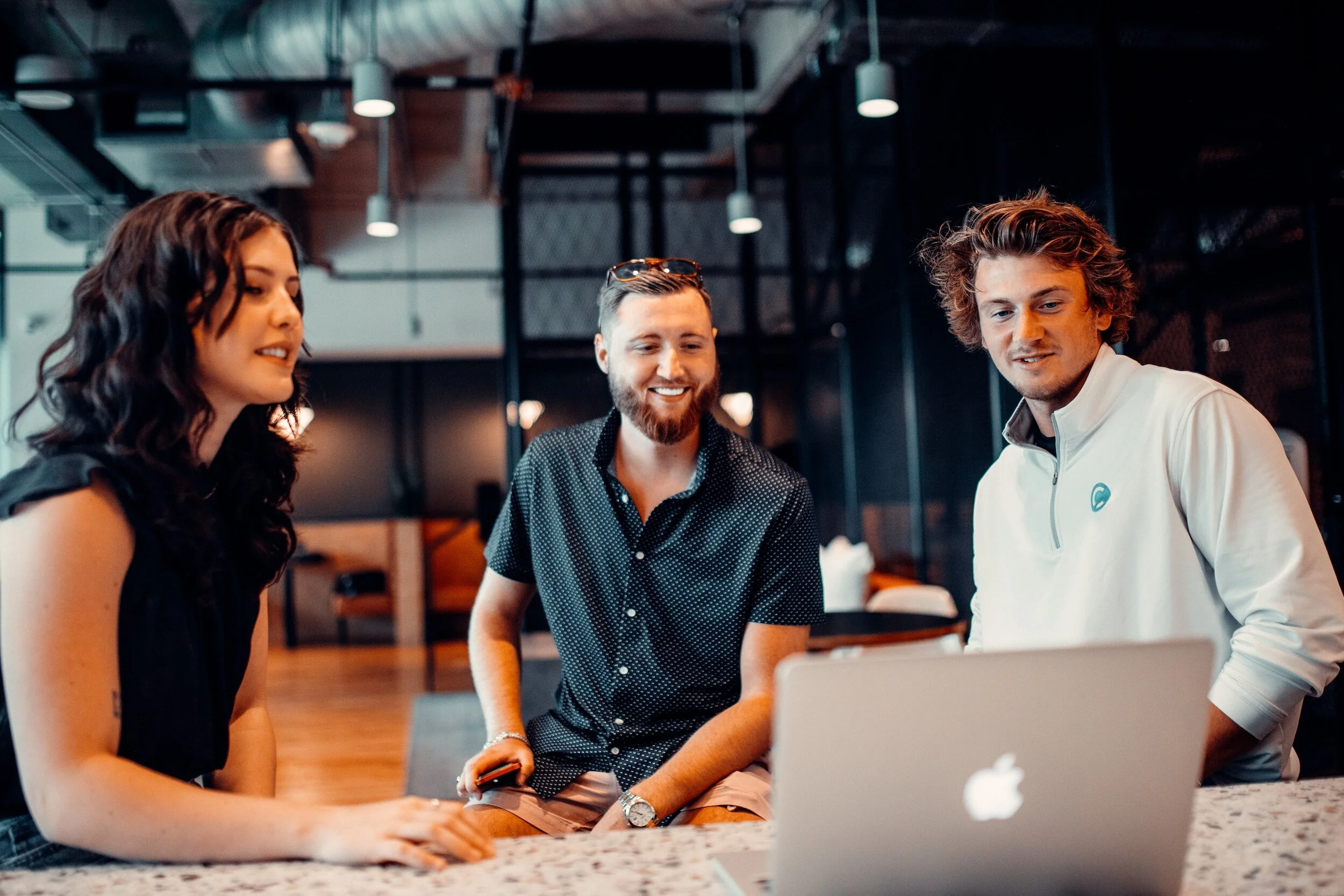 Three young adults, two men and one woman, gathered around a laptop in a modern, industrial-style co-working space with warm lighting and hanging lights.