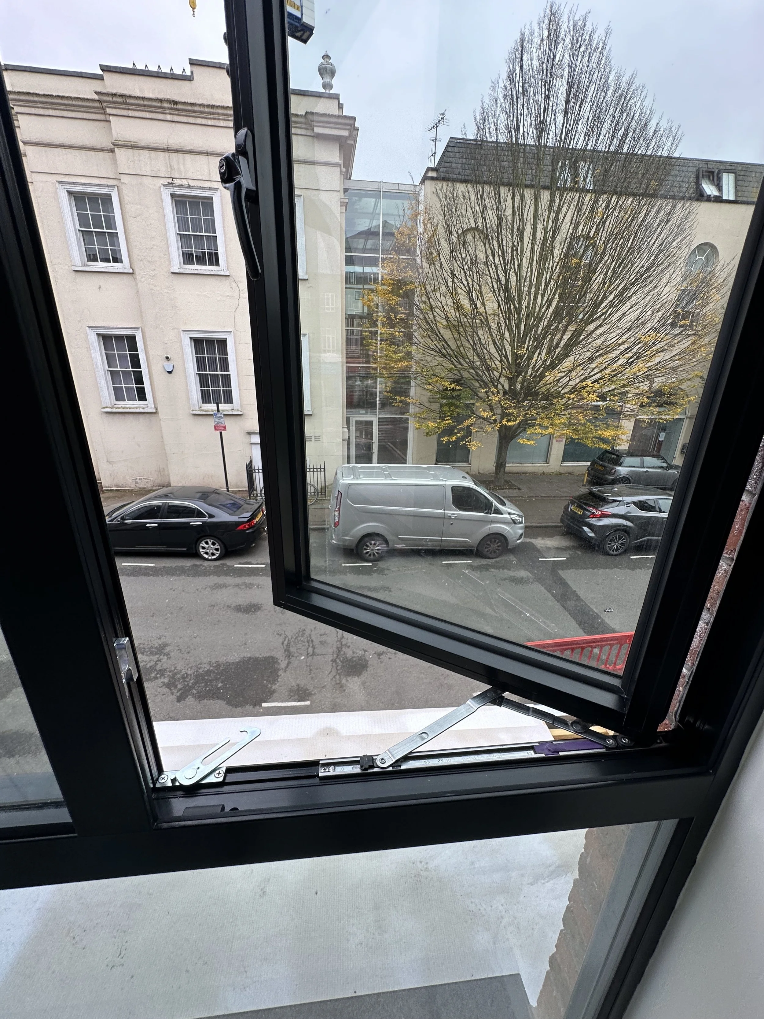 Open window overlooking a city street with parked cars, a tree with yellow leaves, and buildings in the background.