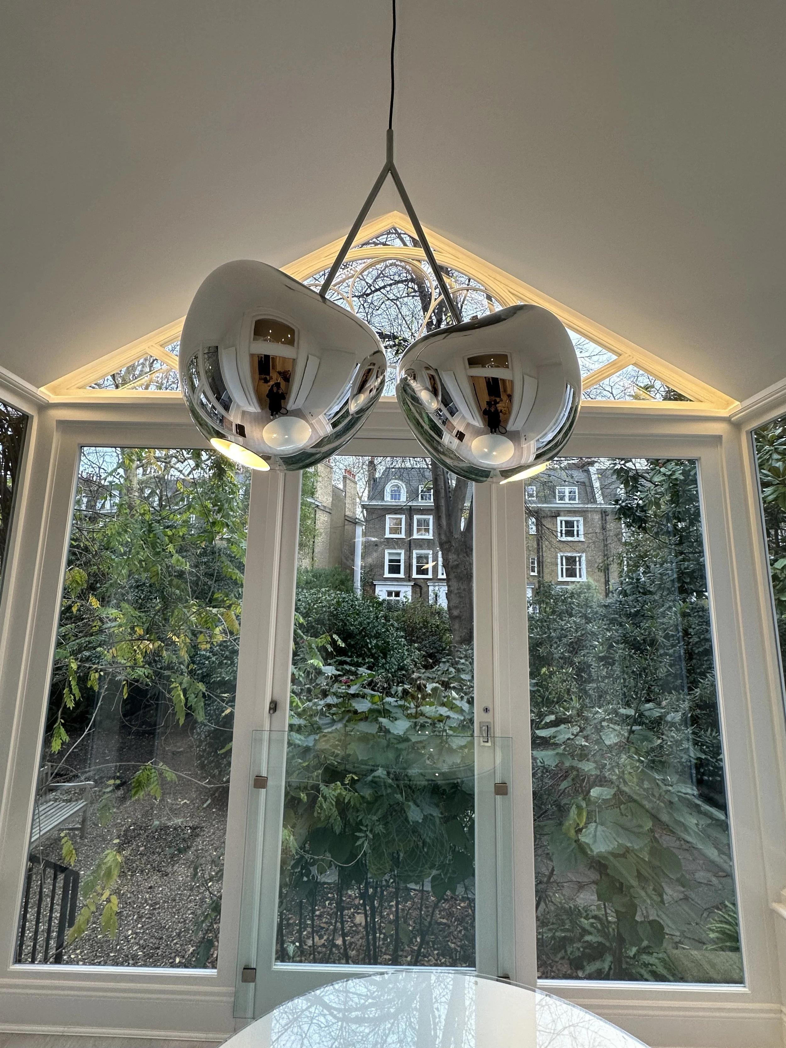 Contemporary dining room with a large window view of greenery and neighboring buildings, featuring a modern chrome hanging light fixture with two spherical reflective bulbs.