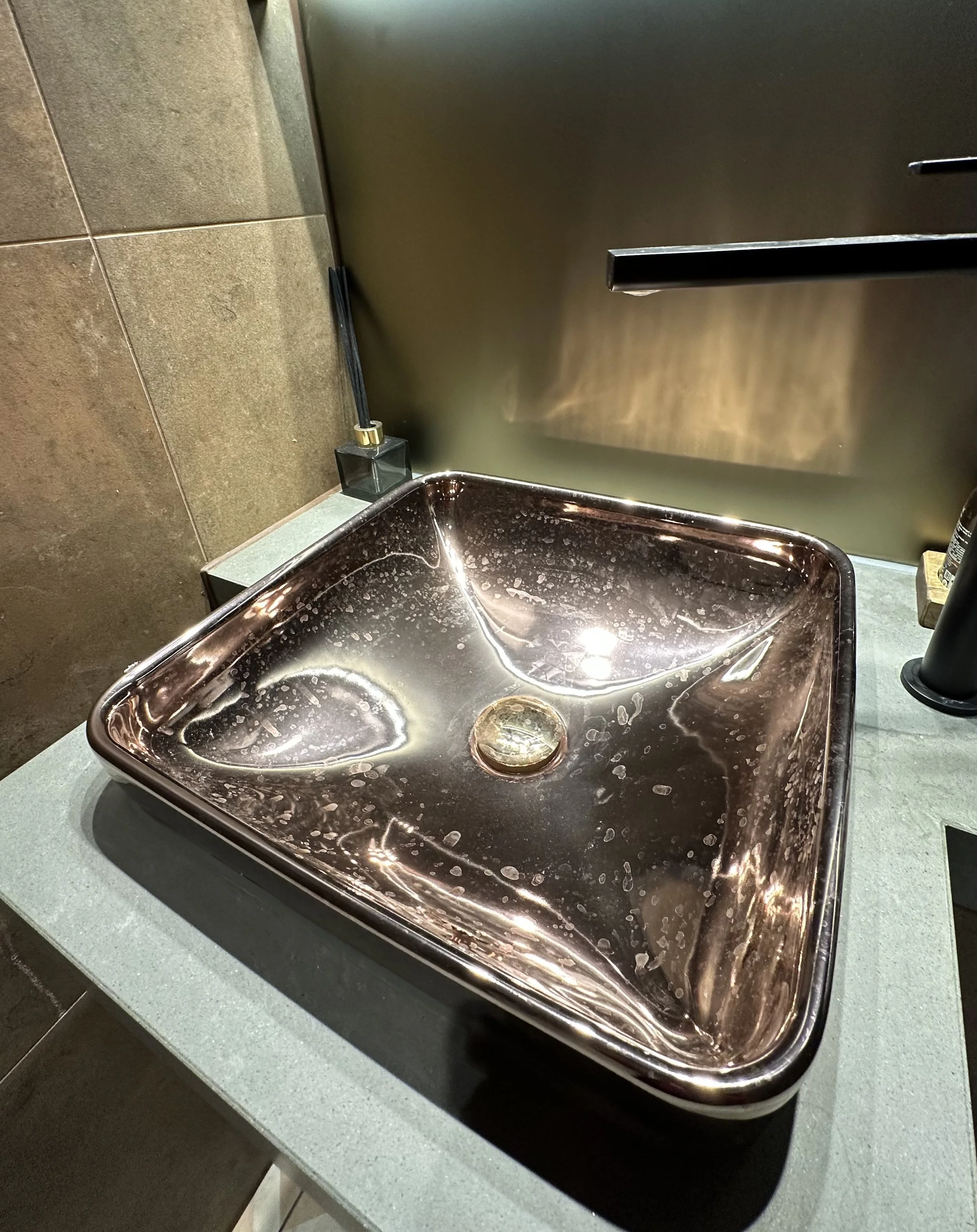A shiny, metallic, square-shaped bathroom sink with soap and water stains, mounted on a green countertop. The sink is positioned under a dark-colored paper towel dispenser or hand dryer in a bathroom setting.