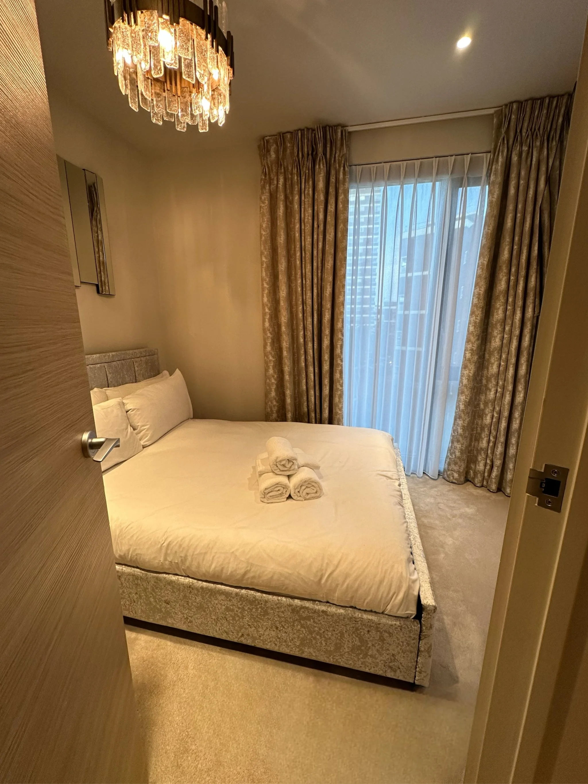 A cozy hotel bedroom with a neatly made bed, three rolled towels on top, beige and gold curtains, a large window with sheer curtains revealing city buildings outside, and a ceiling chandelier.