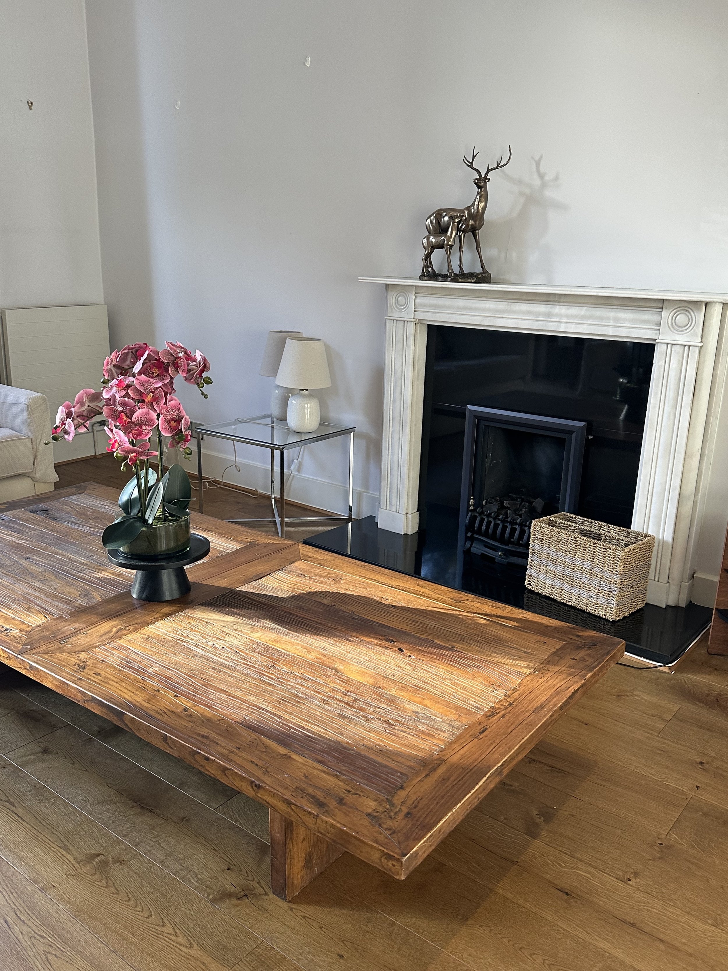 Living room with a large wooden coffee table, a pink orchid in a dark pot, a glass side table with two lamps, a fireplace with a decorative deer sculpture on the mantel, and a woven basket on the hearth.