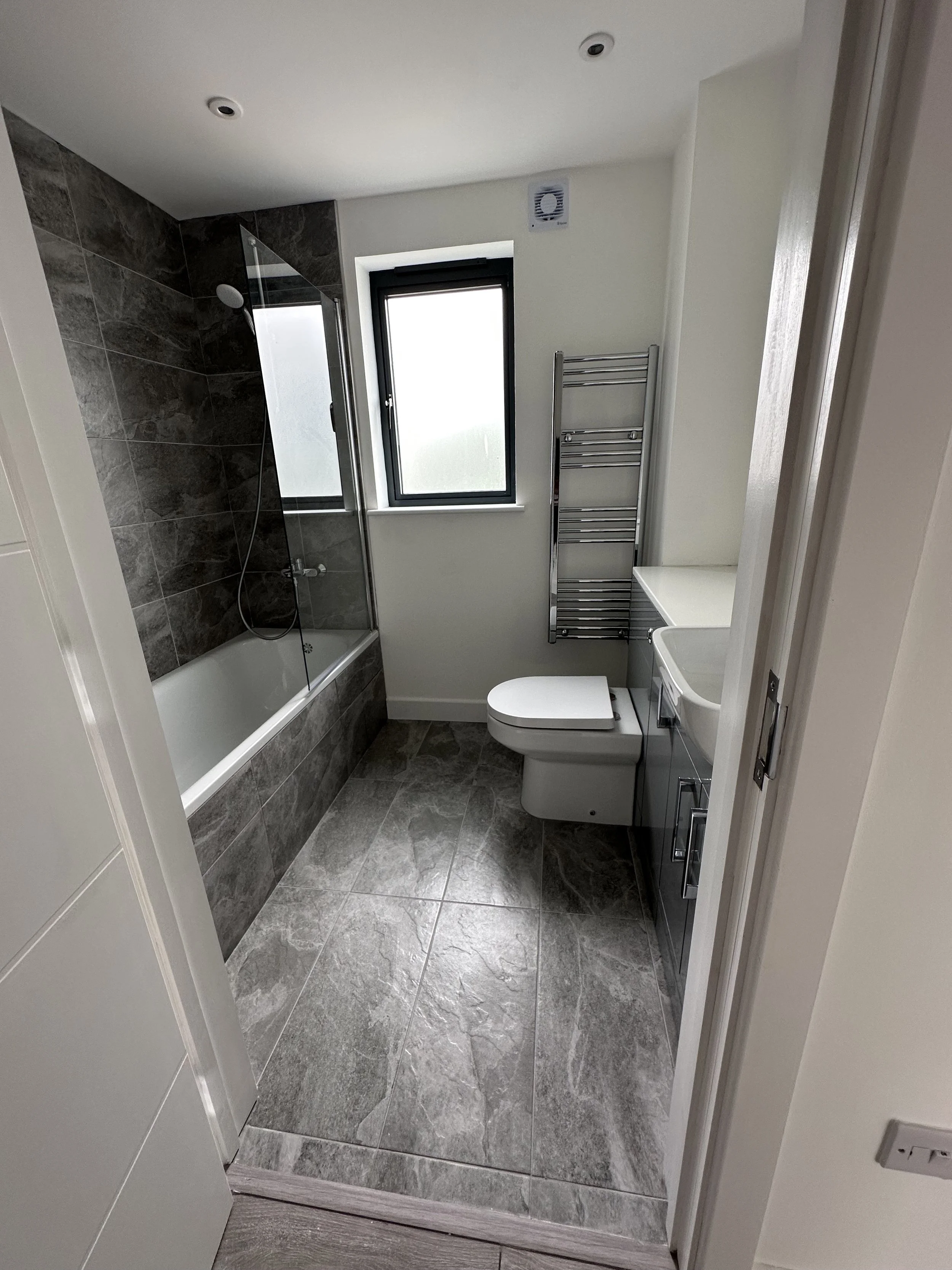 A modern bathroom with gray marble floor tiles, a towel radiator, a small sink with a cabinet, a toilet, a window, and a shower with a glass partition.