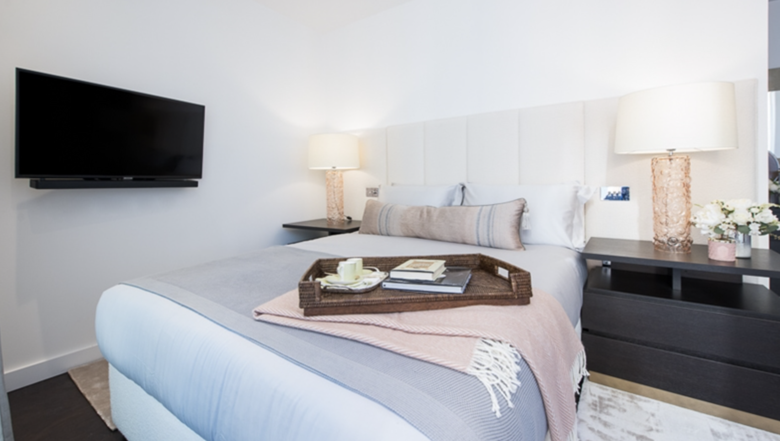 Modern hotel bedroom with a large bed, bedside tables with lamps, a wall-mounted TV, and a tray with books and candles on the bed.