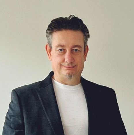 A man with medium-length, slightly wavy brown hair and a goatee, smiling, wearing a black collared shirt.