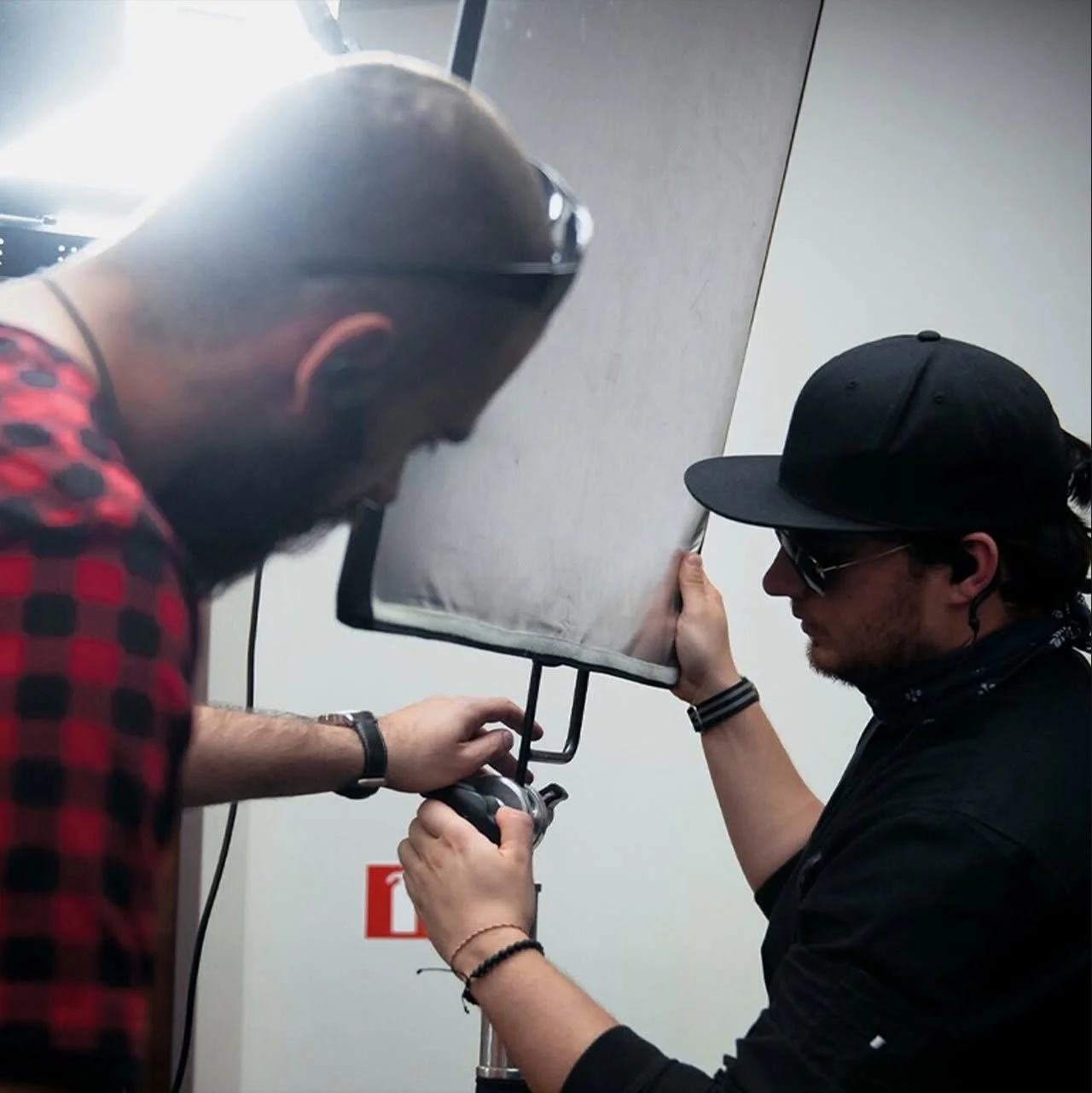 Two men working on a piece of equipment, one wearing a black cap and sunglasses, the other in a helmet and red plaid shirt, in an indoor setting.