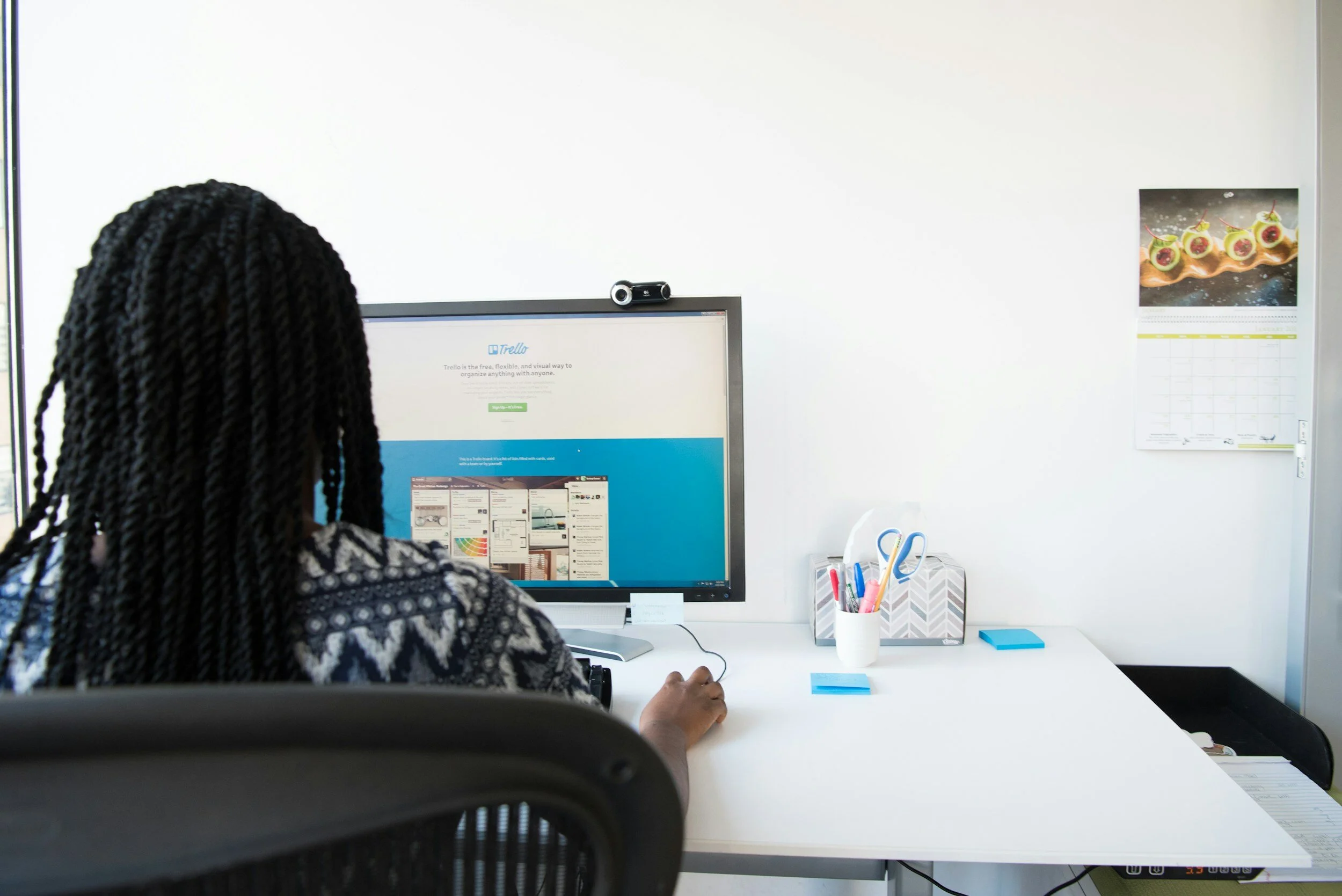 A woman with braided hair working at a desk with a computer monitor in an office. The desk has office supplies, a calendar, and sticky notes. There is a window on the left and a poster calendar on the wall on the right.