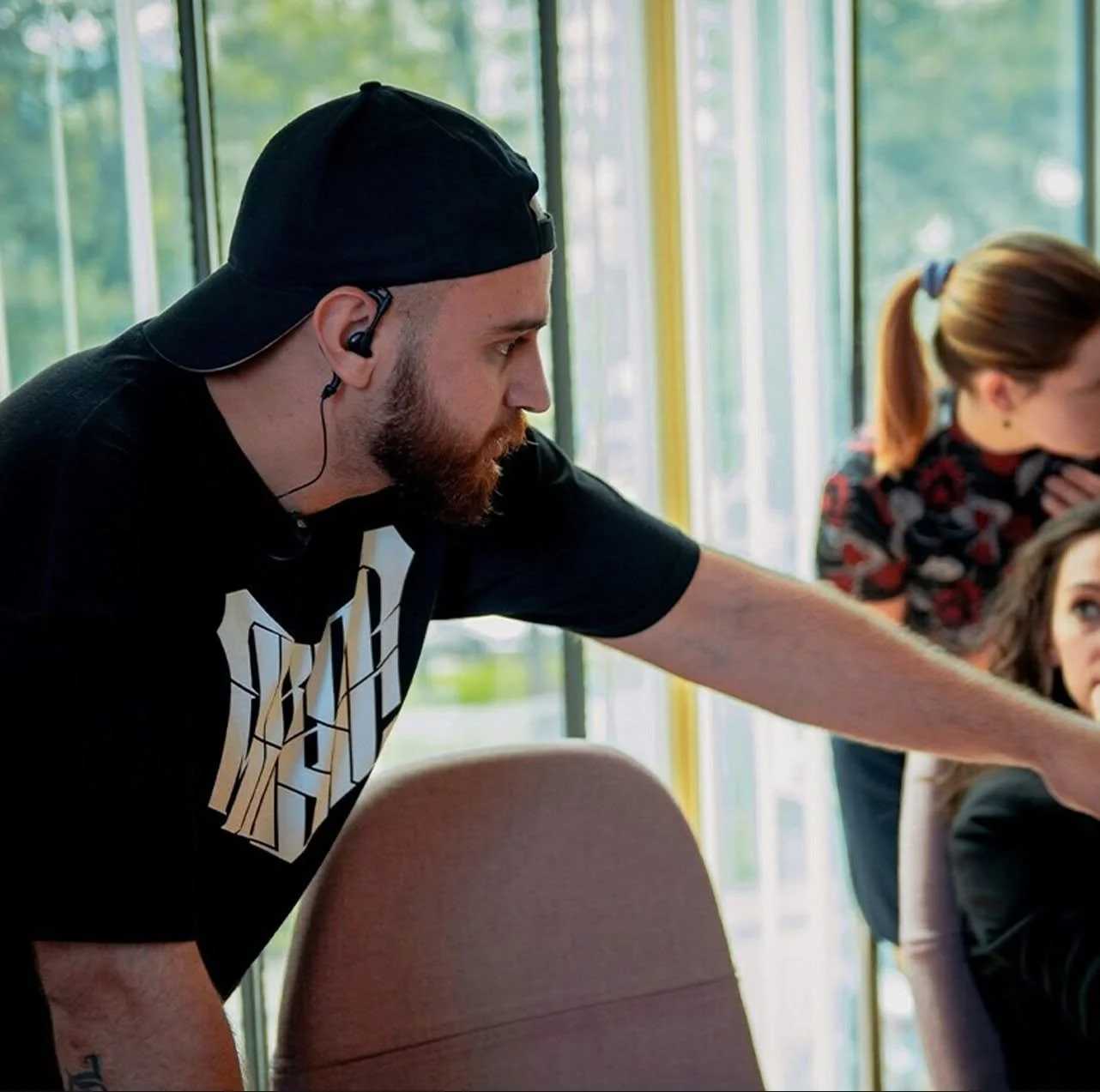 A man with a beard wearing a black cap and earbud, leaning forward and pointing at something, sitting next to a woman with brown hair tied back, in a room with large windows and green outdoor scenery.