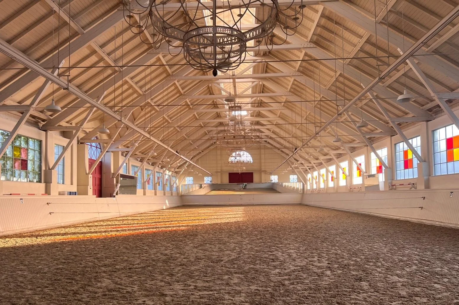 Leerer Reitplatz im Innenraum mit Holzdecke, Fenstern mit buntem Glas, großen alten Kronleuchtern und Sandboden.