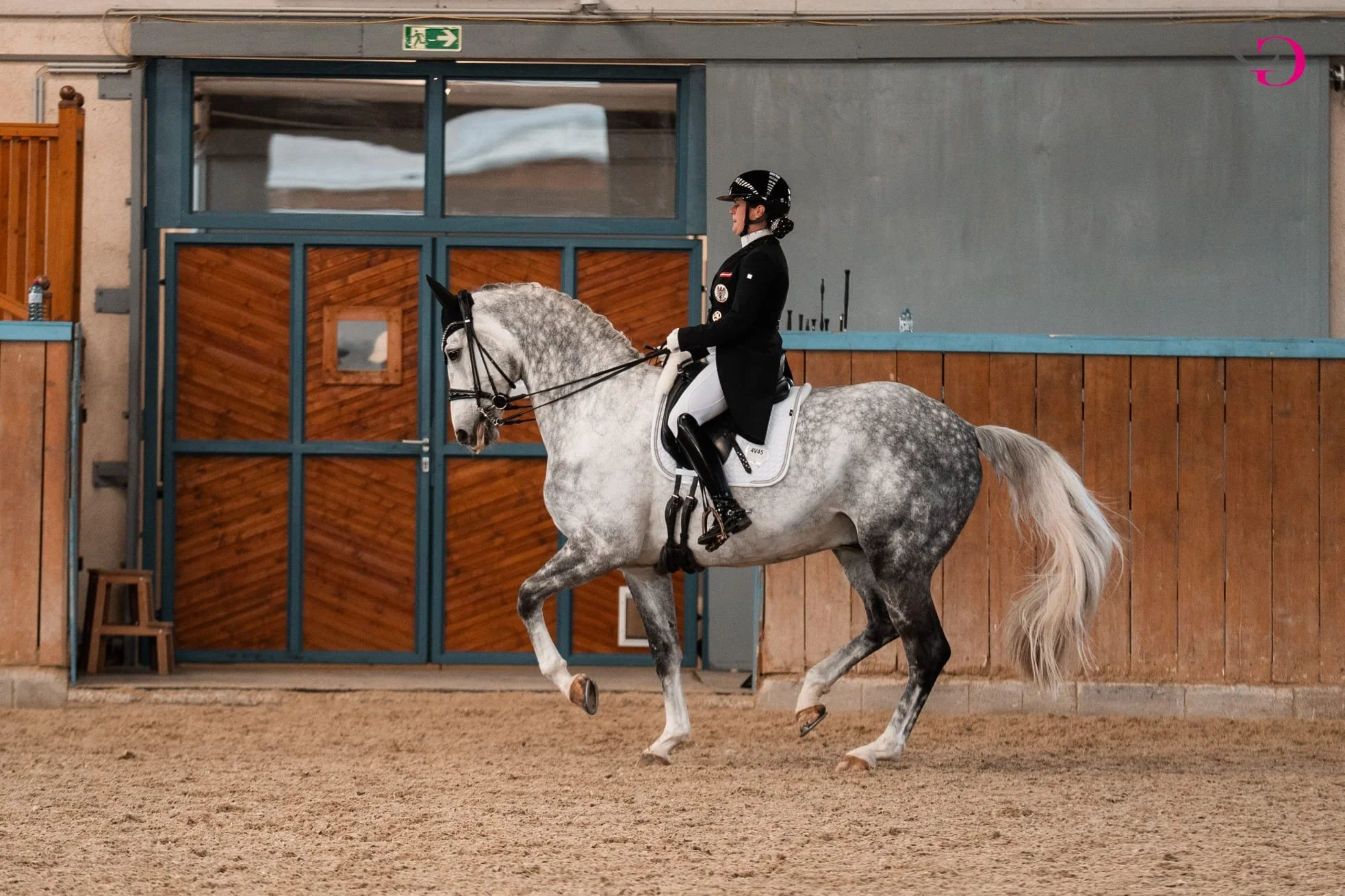 Reiterin auf grauem Pferd in einer Reithalle auf weißem Sandboden. Die Reiterin trägt eine schwarze Jacke, weiße Reithose, schwarzen Stiefel und einen schwarzen Helm. Im Hintergrund eine Tür mit Holz und blaue Rahmen, oben ein grünes Notausgangsschil