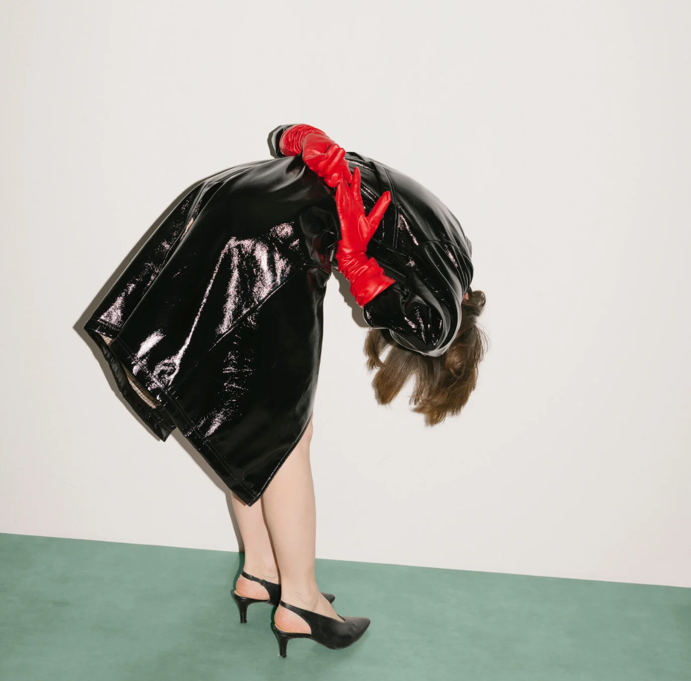 Une femme portant une veste en cuir noir brillant, des chaussures à talons noires, et des gants rouges, tandis qu'elle se penche en avant contre un mur blanc, avec ses cheveux bruns visibles.