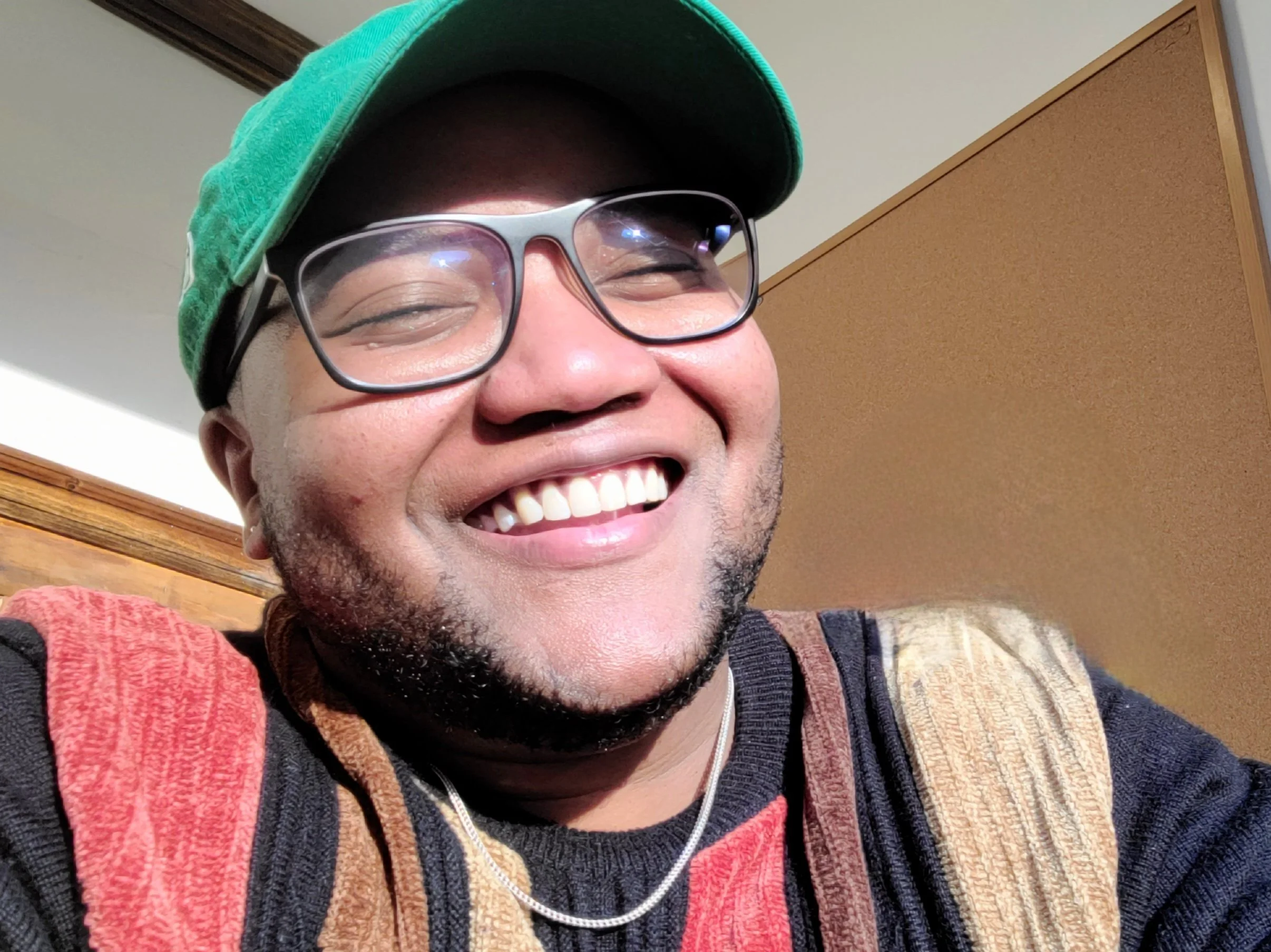 Kaden, a black man in a green hat and striped sweater, glasses, and chain smiling really big in the sunlight.