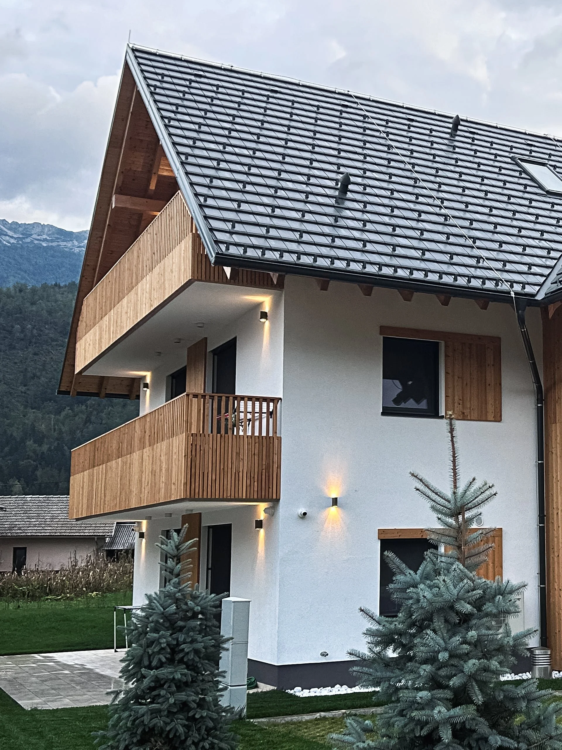 A modern white house with wooden balconies, black window frames, and a dark gray tiled roof, situated in a mountainous area of Bohinj.