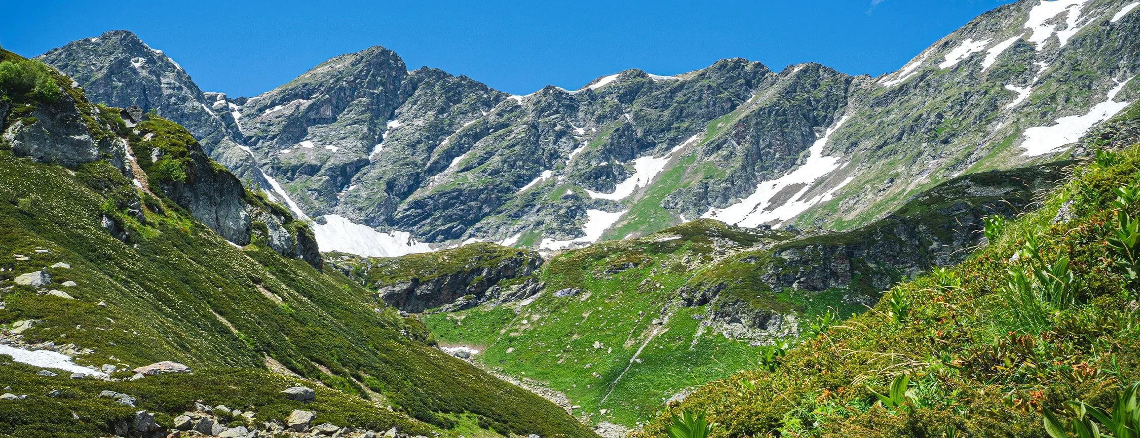 Mountain landscape with rocky peaks, some patches of snow, and green vegetation under a clear blue sky.
