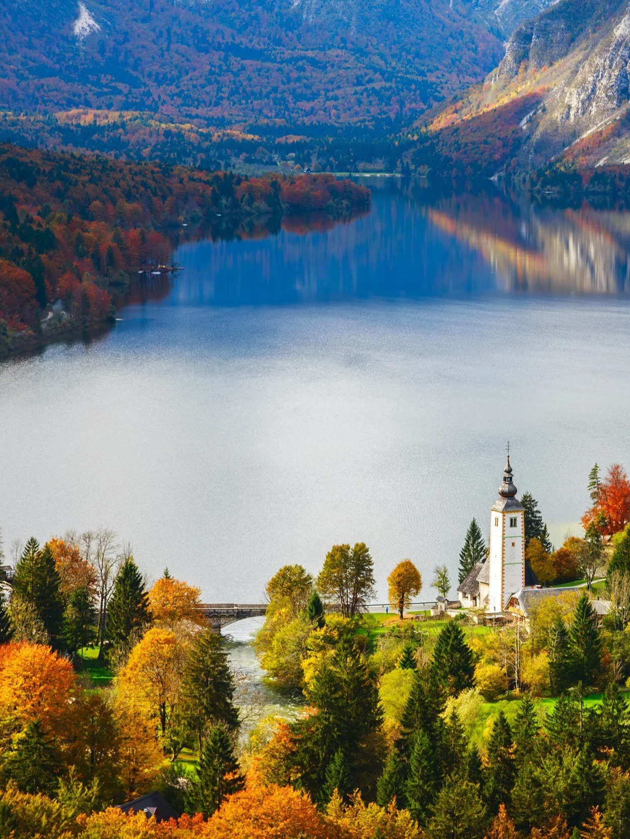 Autumn landscape in Bohinj with a lake, colorful trees, and a church with a tall steeple near the water.